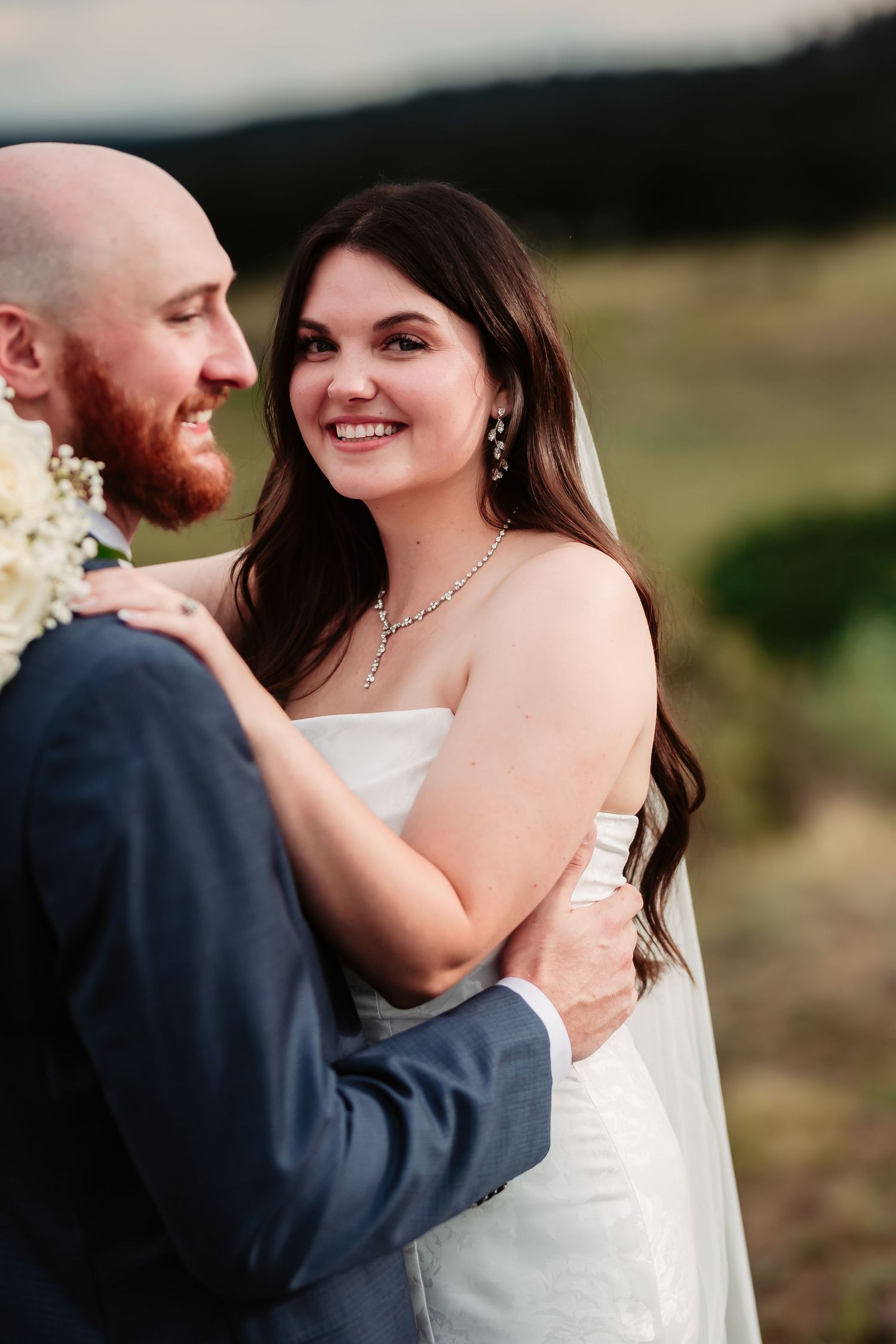 A joyful wedding couple embraces outdoors with mountains in the background during their romantic photoshoot.