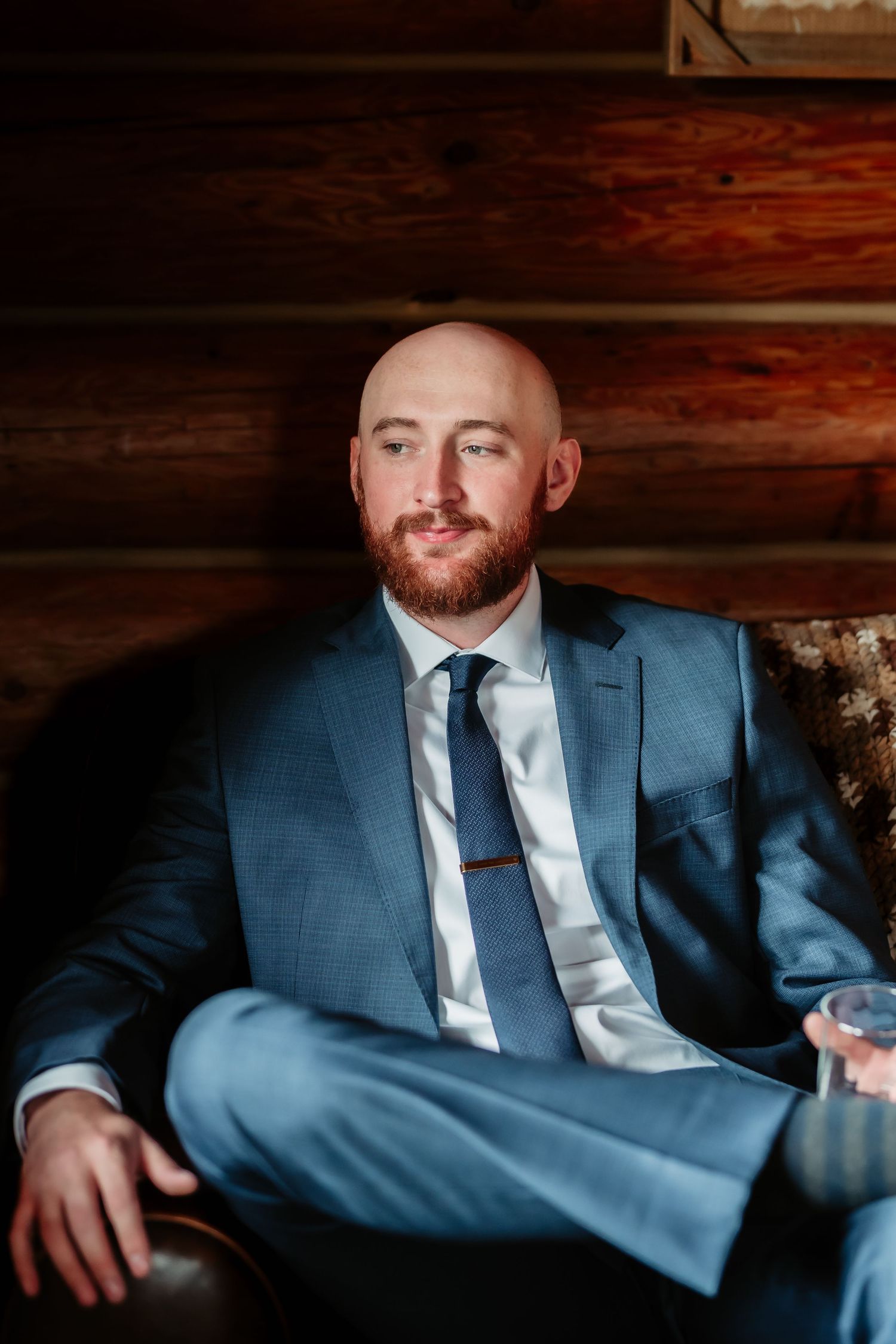 A groom in a blue suit and silver tie sits in a rustic wooden cabin setting.