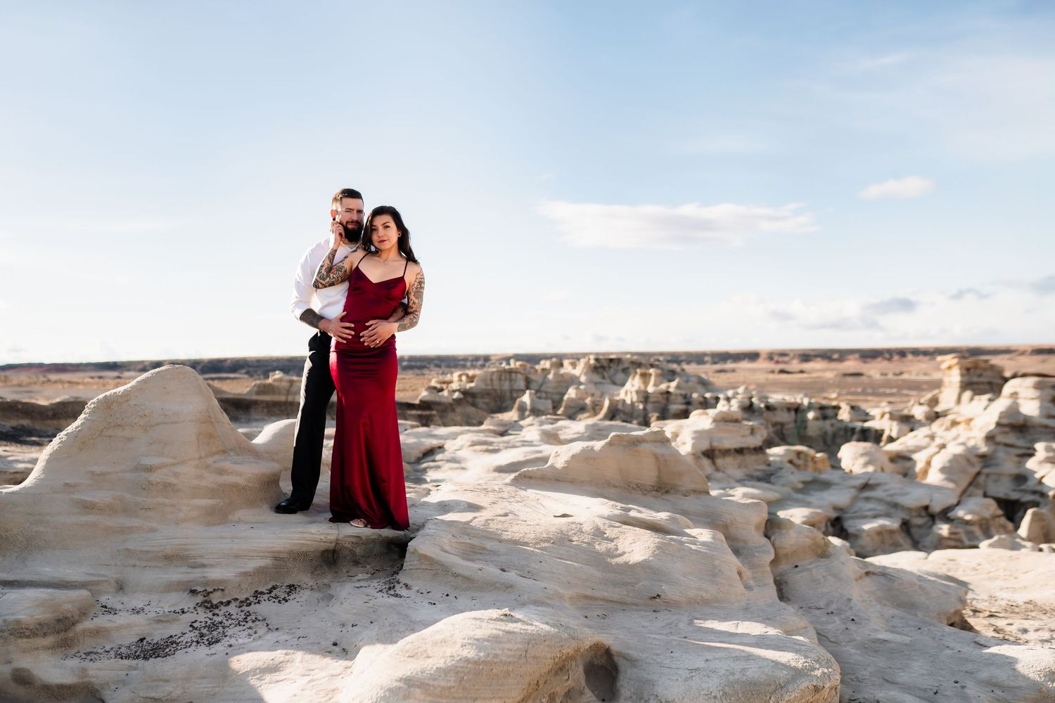 A couple in formal wear embraces on rocky terrain with desert landscape behind them.