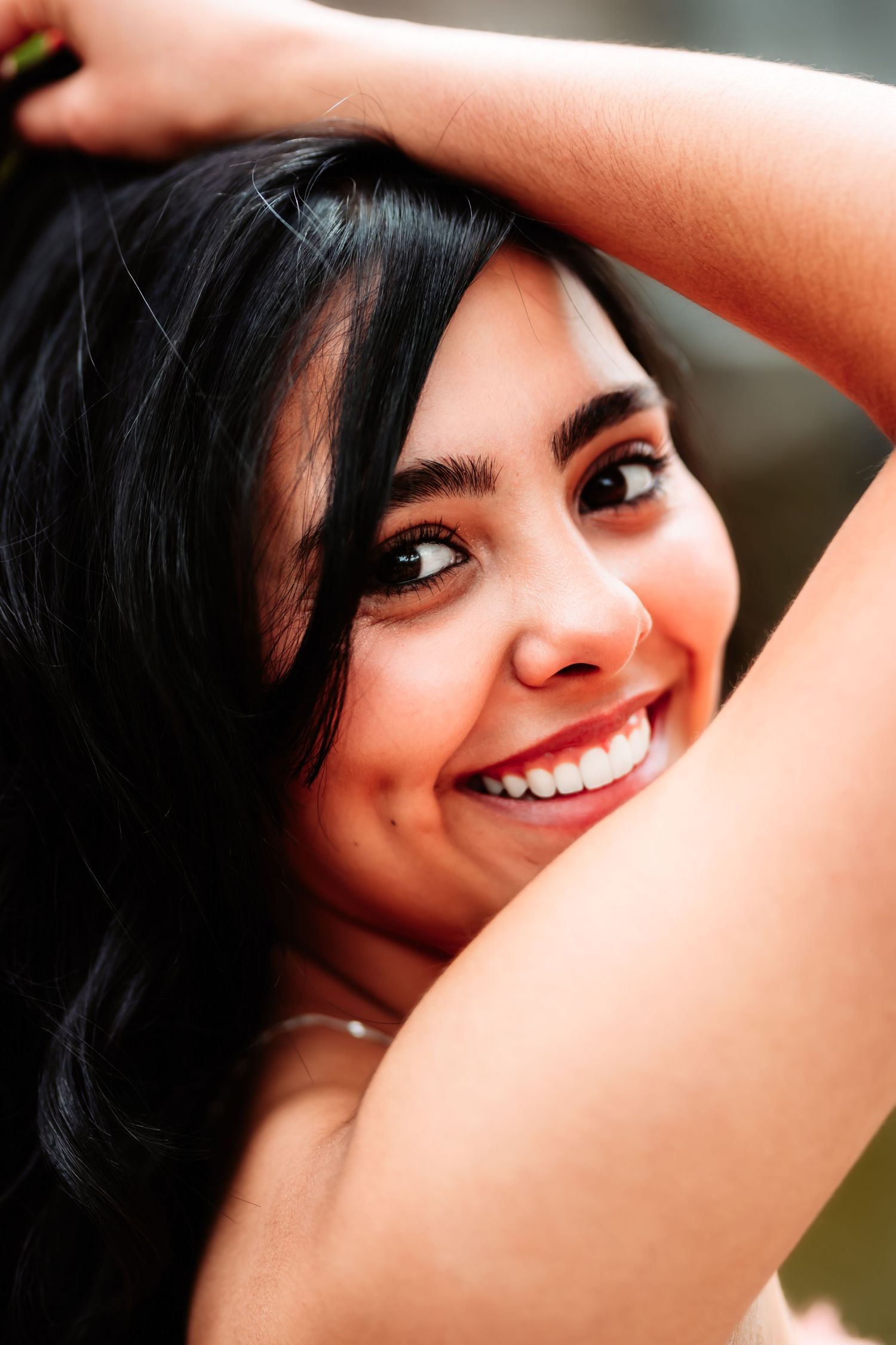 Closeup portrait showing a bright smile and long dark hair against a blurred natural background.