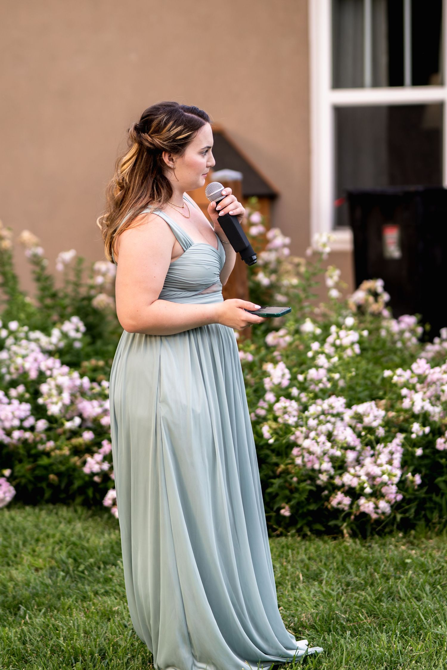 A bridesmaid in a flowing sage green dress speaks into a microphone in a garden setting.