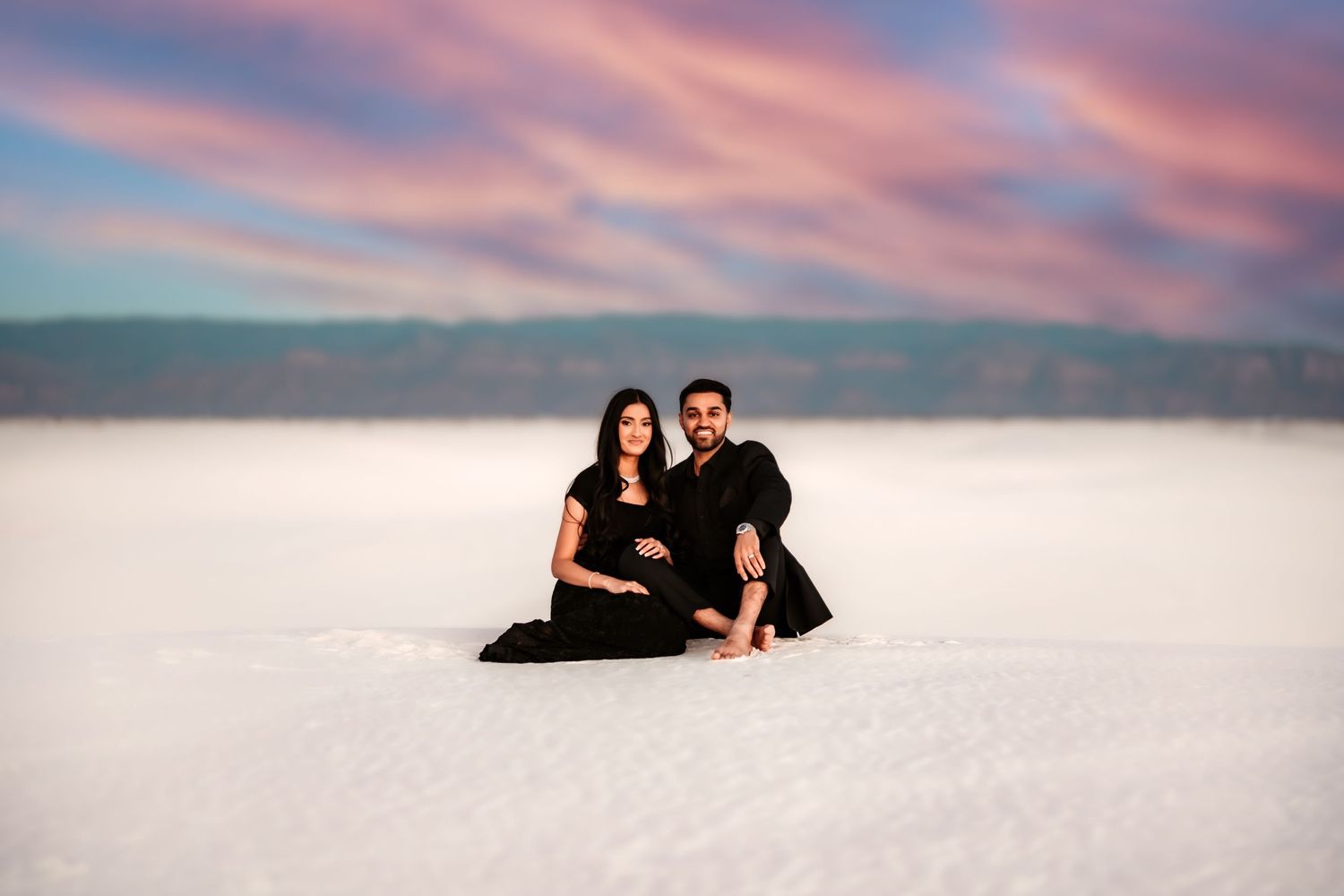 A couple dressed in black sits together on white sand beneath a pink and purple sunset sky.