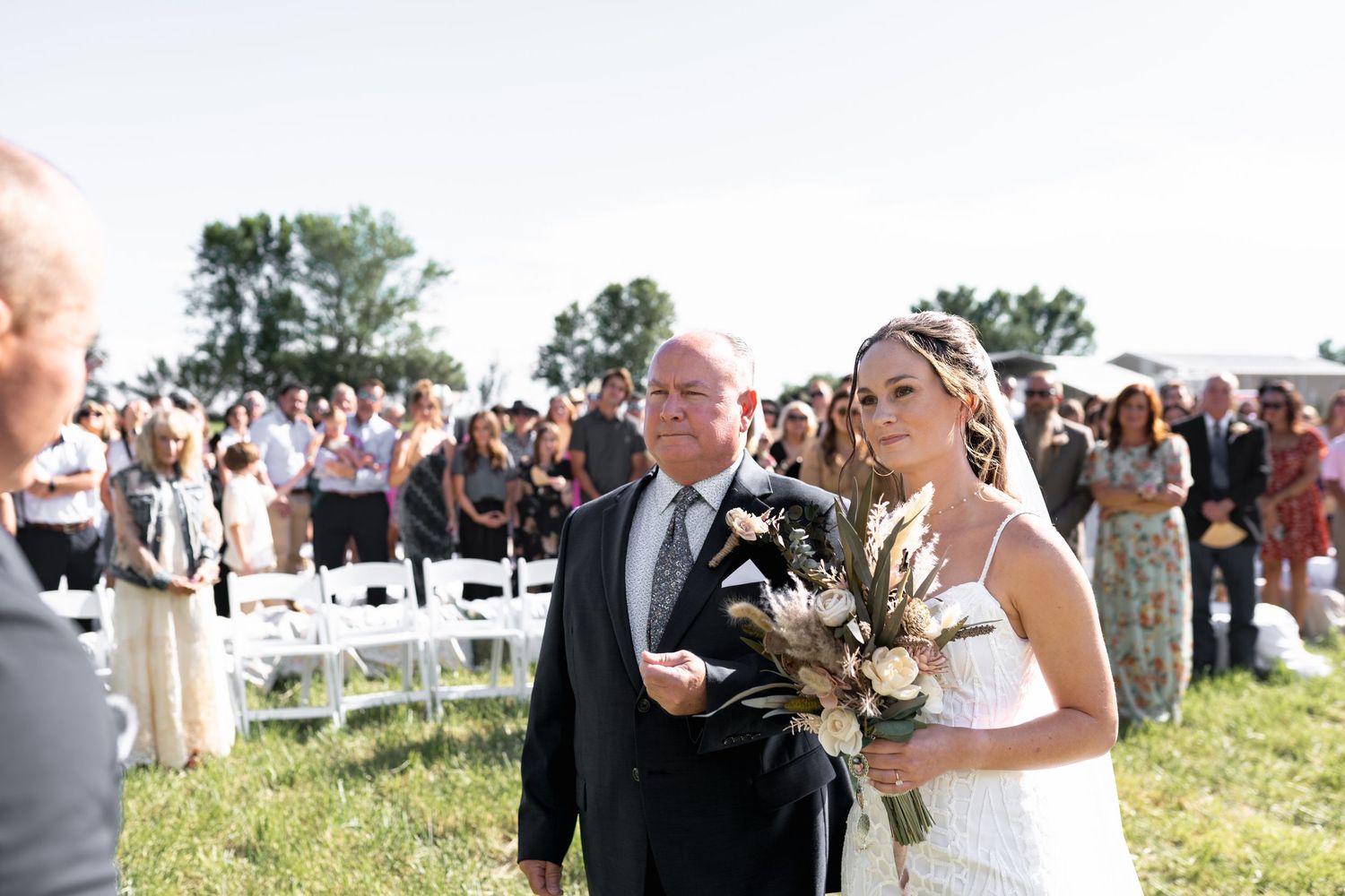Outdoor wedding ceremony with crowd of guests seated in white chairs on grassy field during sunny summer day.