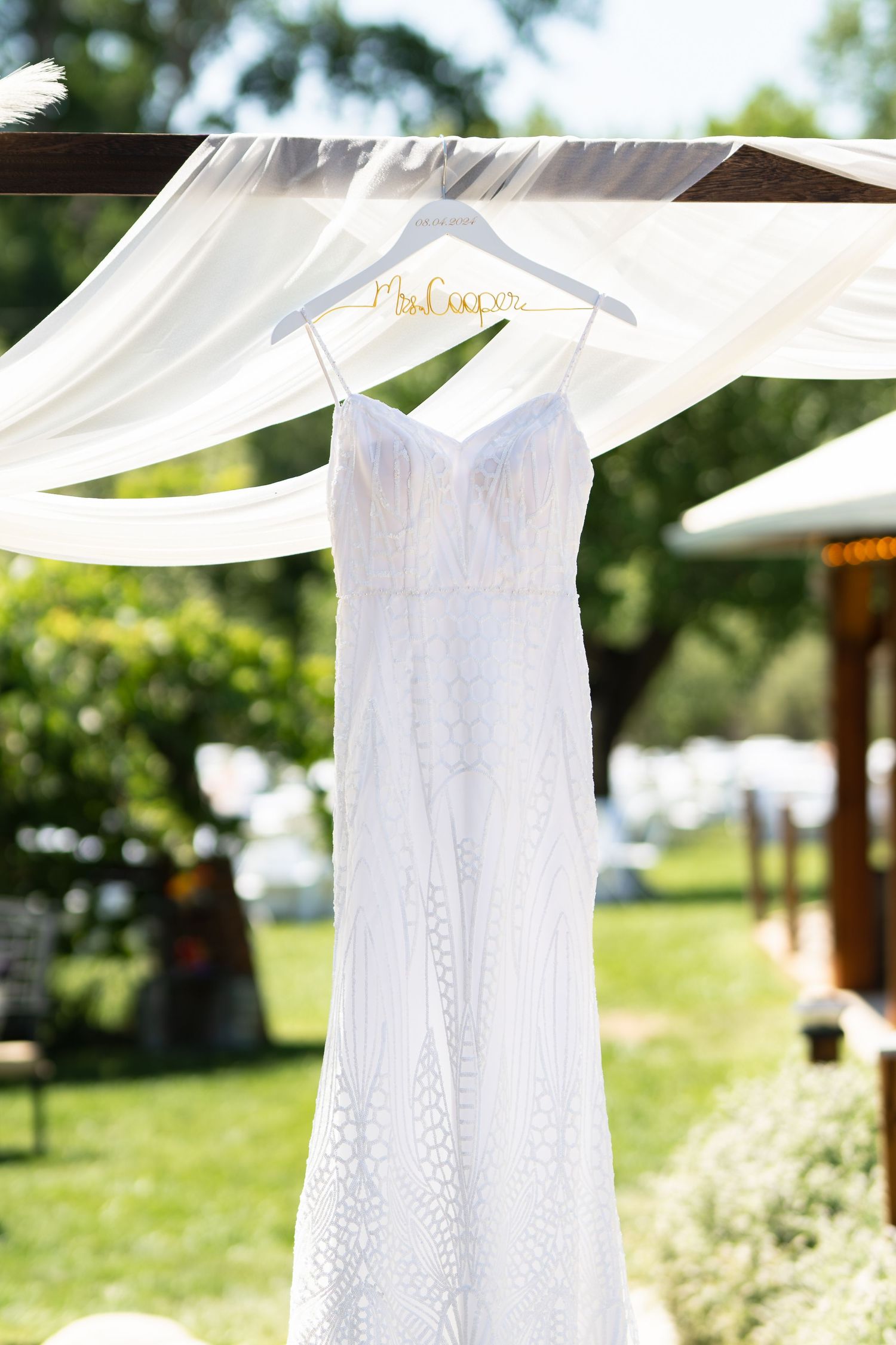 A white lace wedding dress hangs from a wooden hanger against a backdrop of flowing white fabric outdoors.