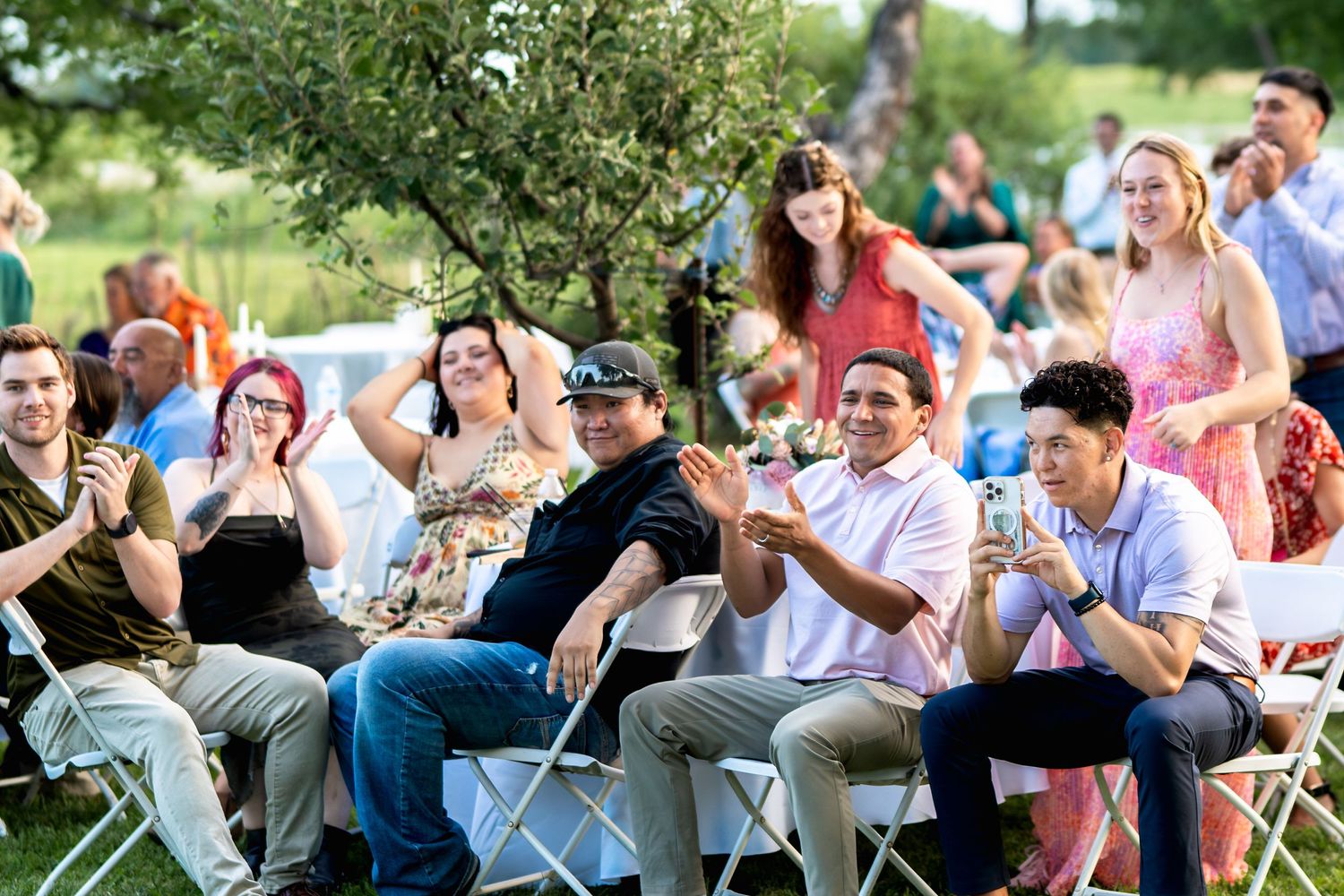 Wedding guests seated in white folding chairs react enthusiastically during an outdoor ceremony.