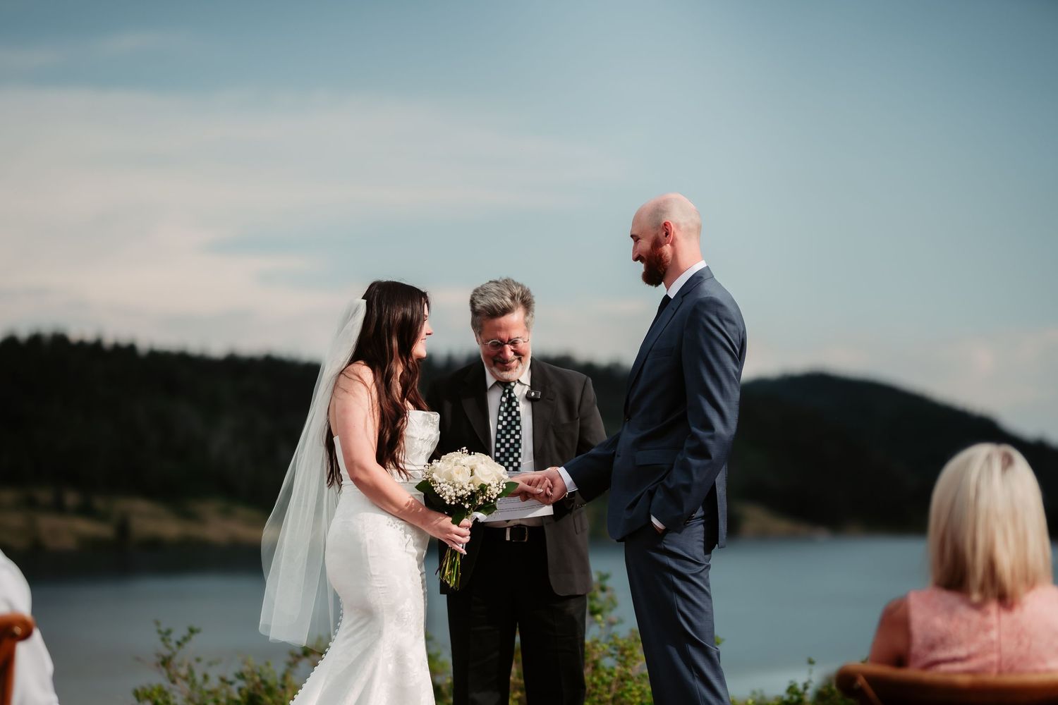 An intimate outdoor wedding ceremony takes place on a clifftop overlooking a serene lake.