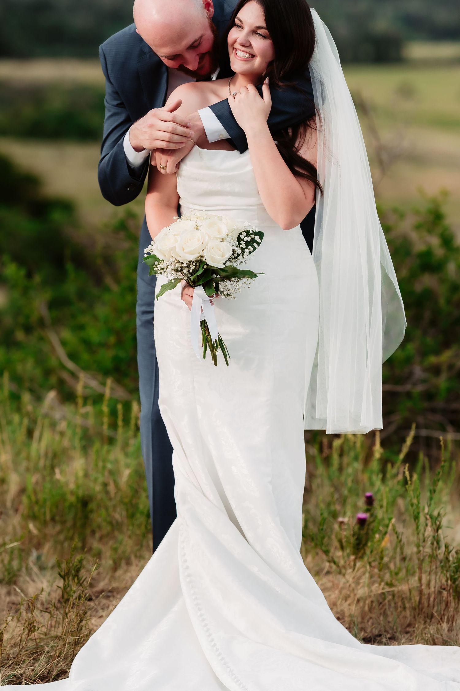 A joyful wedding couple embraces during their outdoor portrait session in a field.