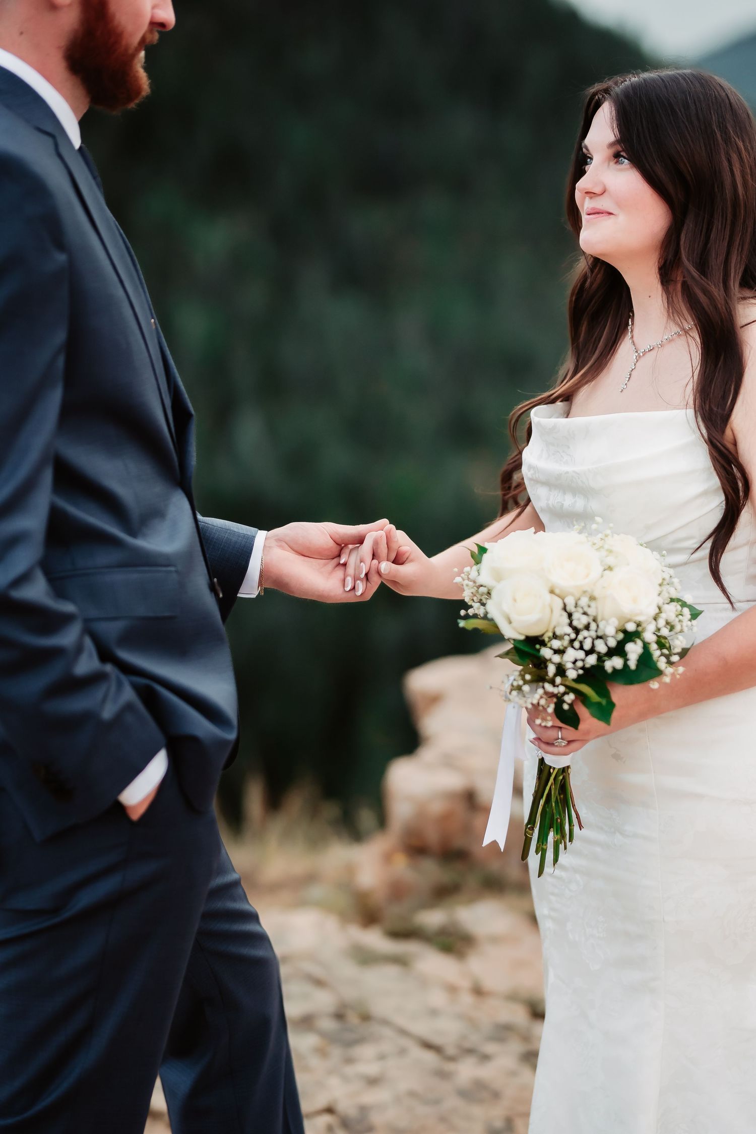 Close-up shot of newlyweds holding hands with white floral bouquet against mountain backdrop.