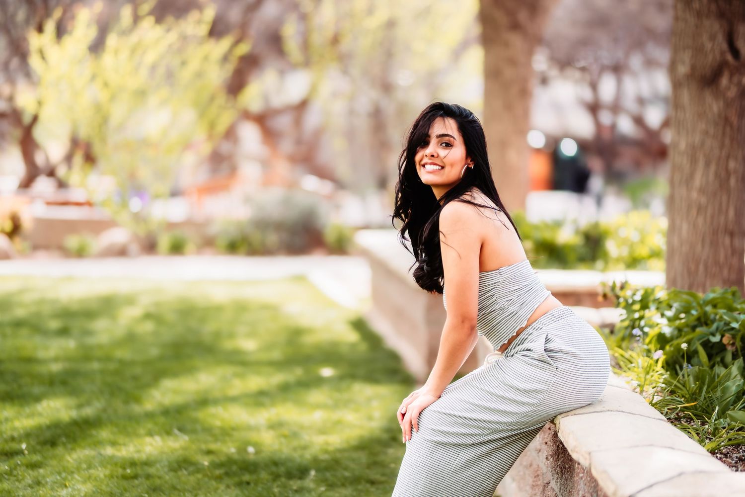 Woman in gray dress poses by stone wall in sunny garden setting.