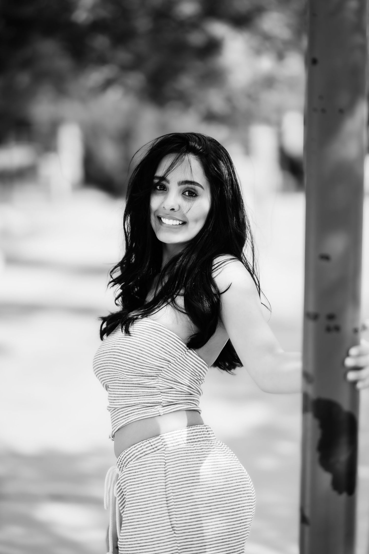 Monochrome photo showing someone in a fitted striped dress posing against a tree trunk outdoors.