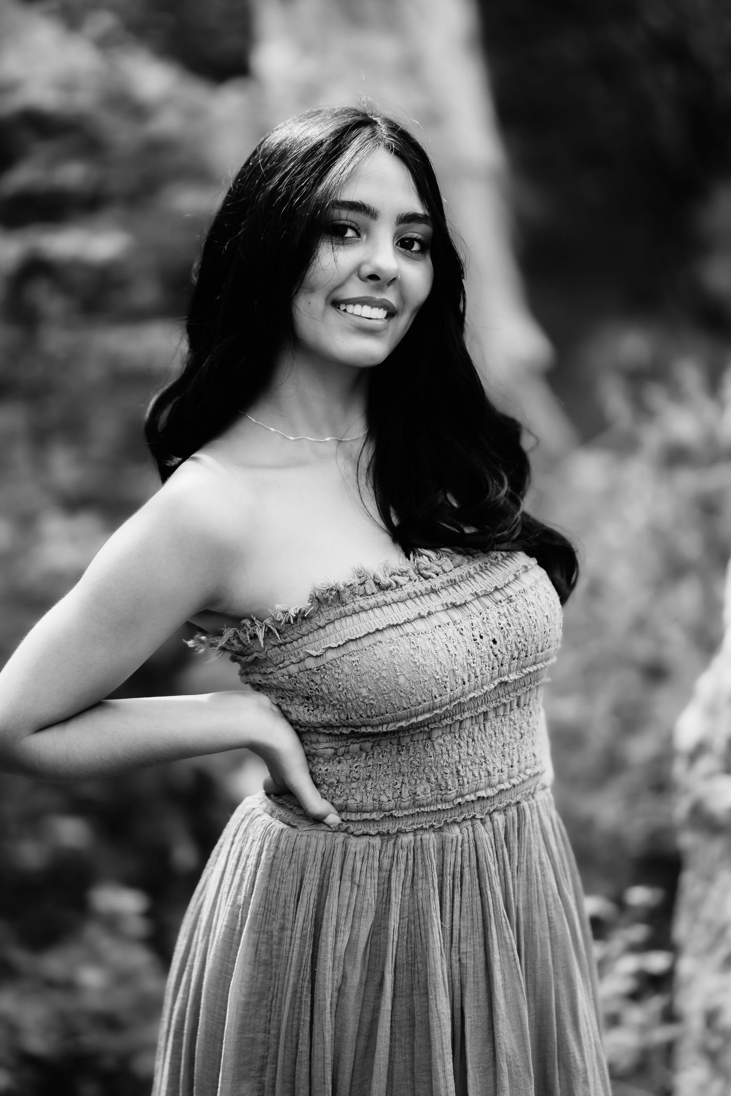 Black and white portrait of woman in smocked dress posing against natural outdoor background.