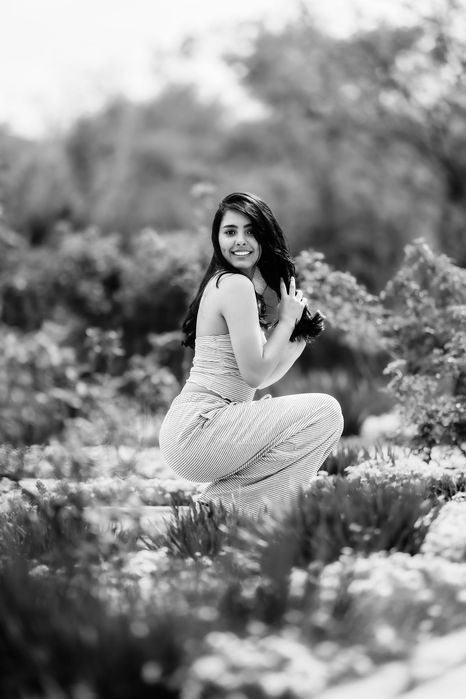 Black and white portrait of a person crouching among flowering plants in a garden setting.