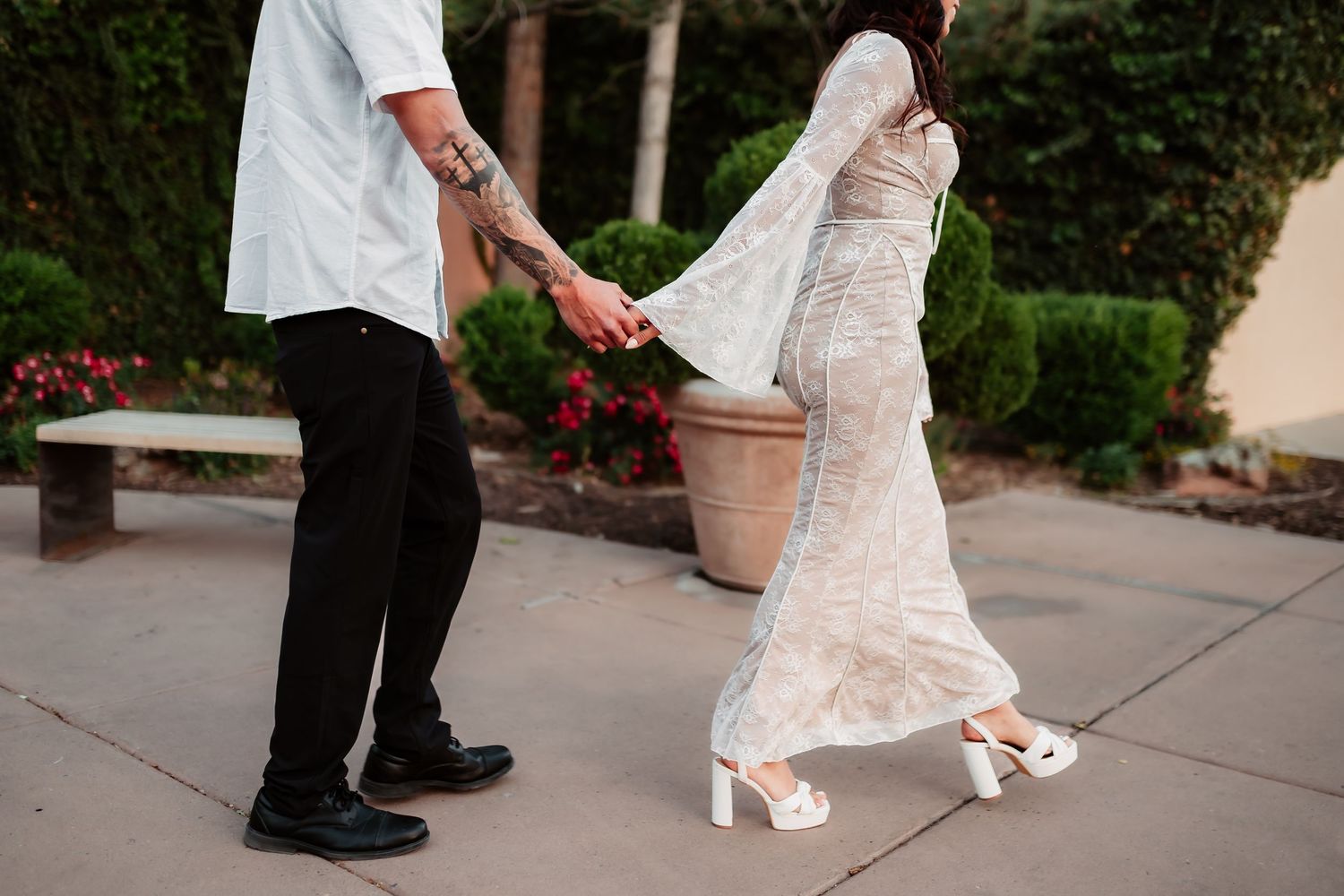 Couple holds hands while walking outdoors near decorative planters and benches.