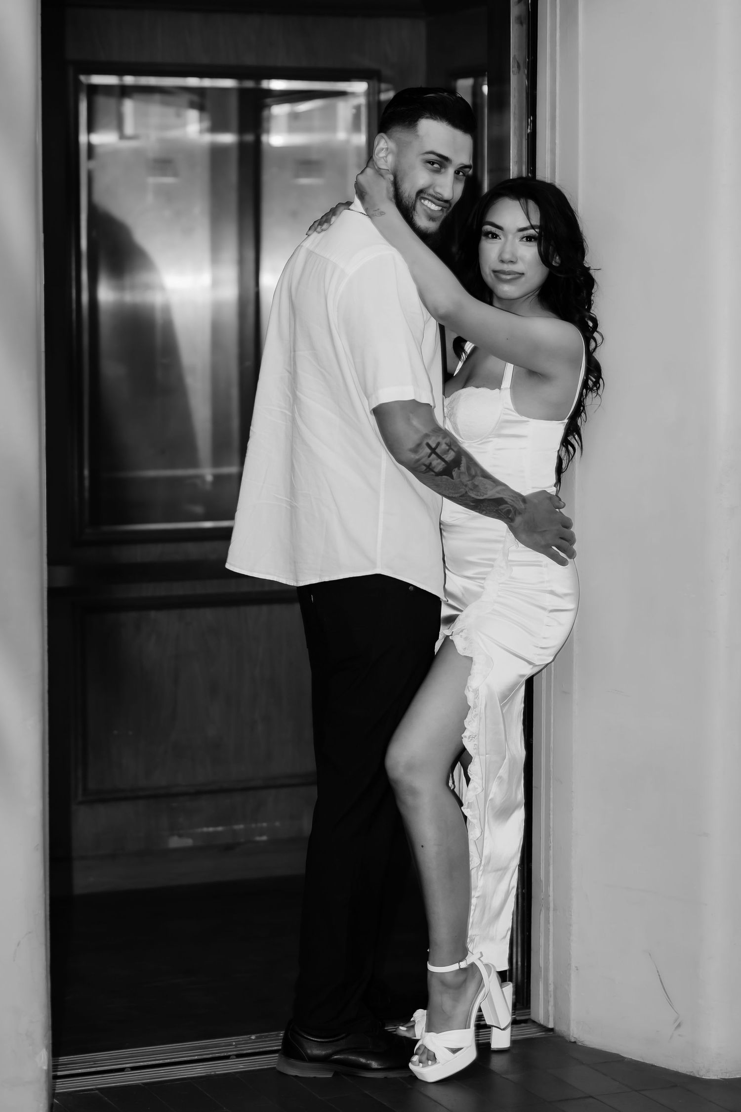 A stylish couple embraces in a black and white portrait by an elevator.