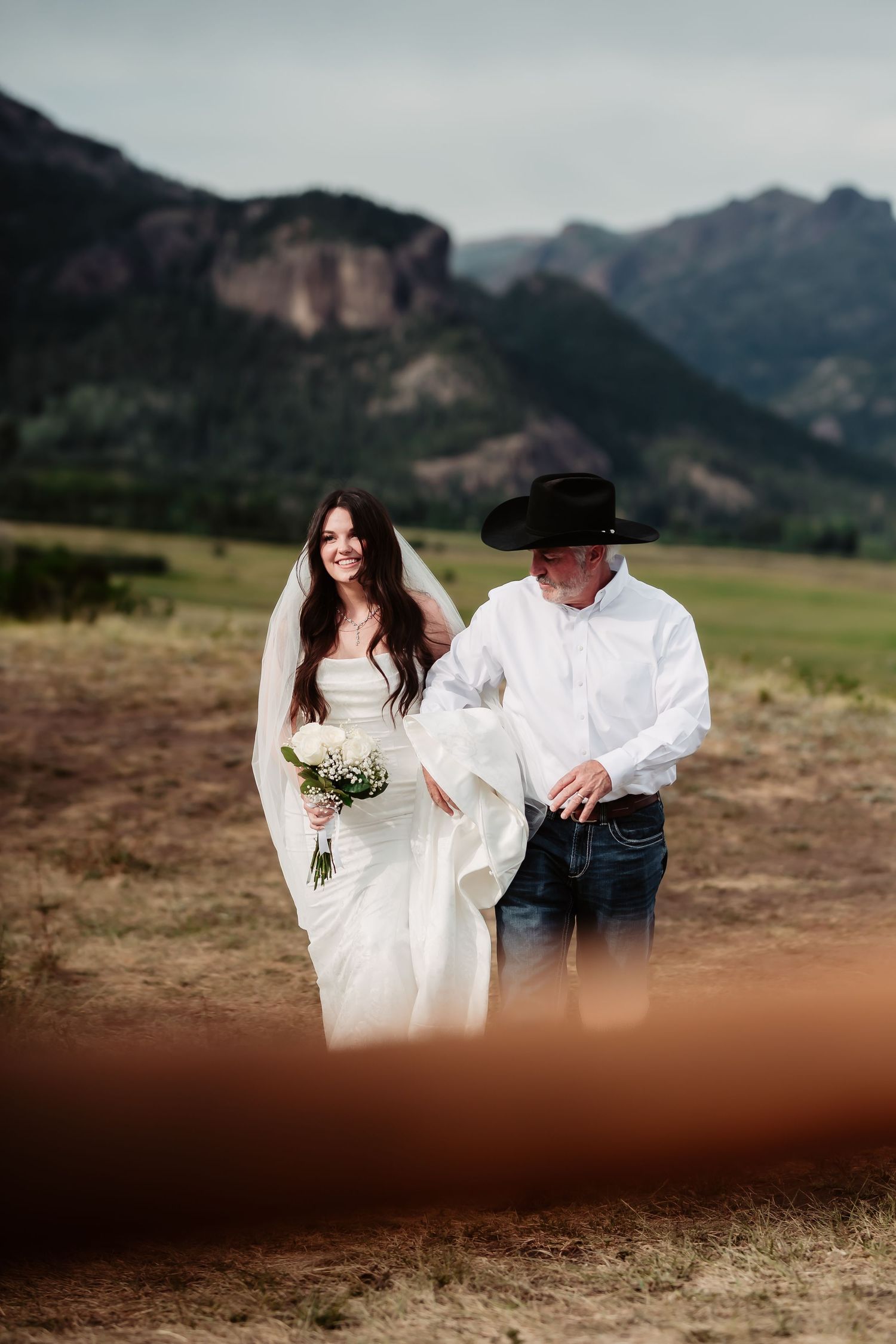 A beautiful outdoor wedding photo with a mountain backdrop showing a bride in white gown walking with a companion.