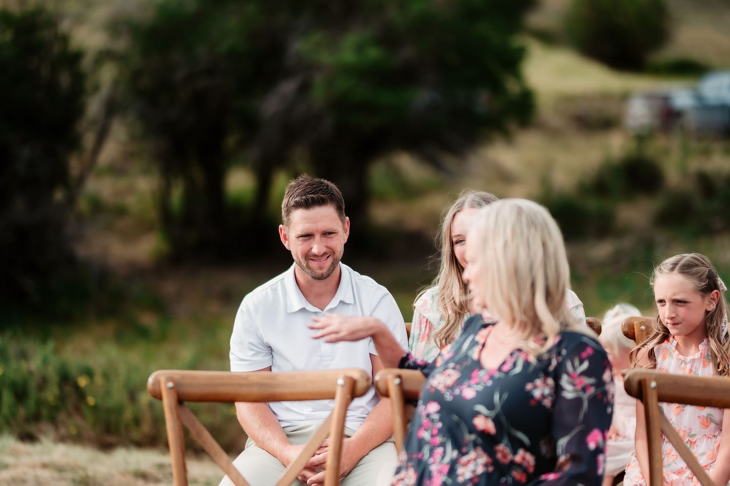 People enjoying an outdoor gathering seated at wooden chairs in a natural setting.