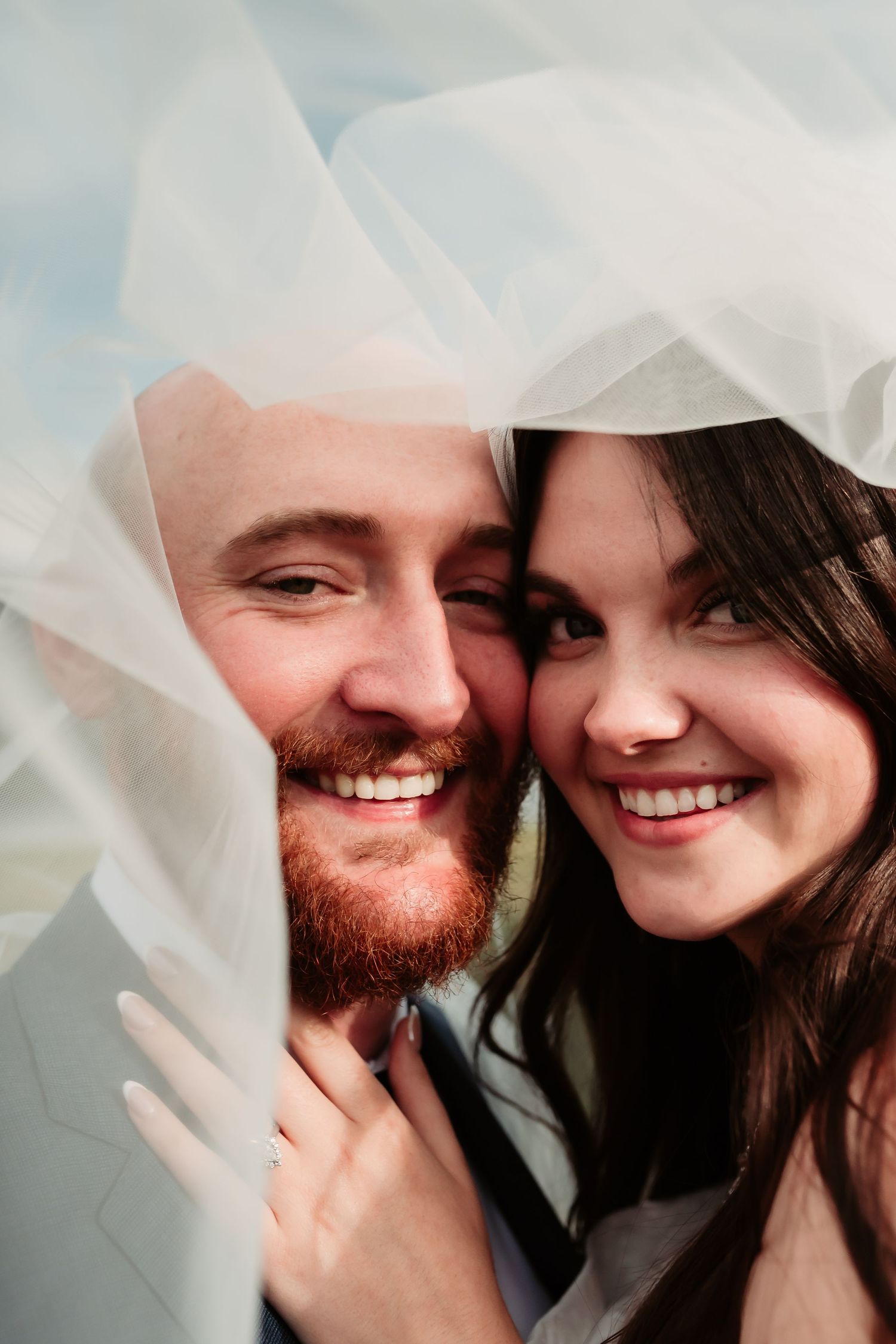 Wedding couple shares intimate moment under flowing white veil against blurred background.