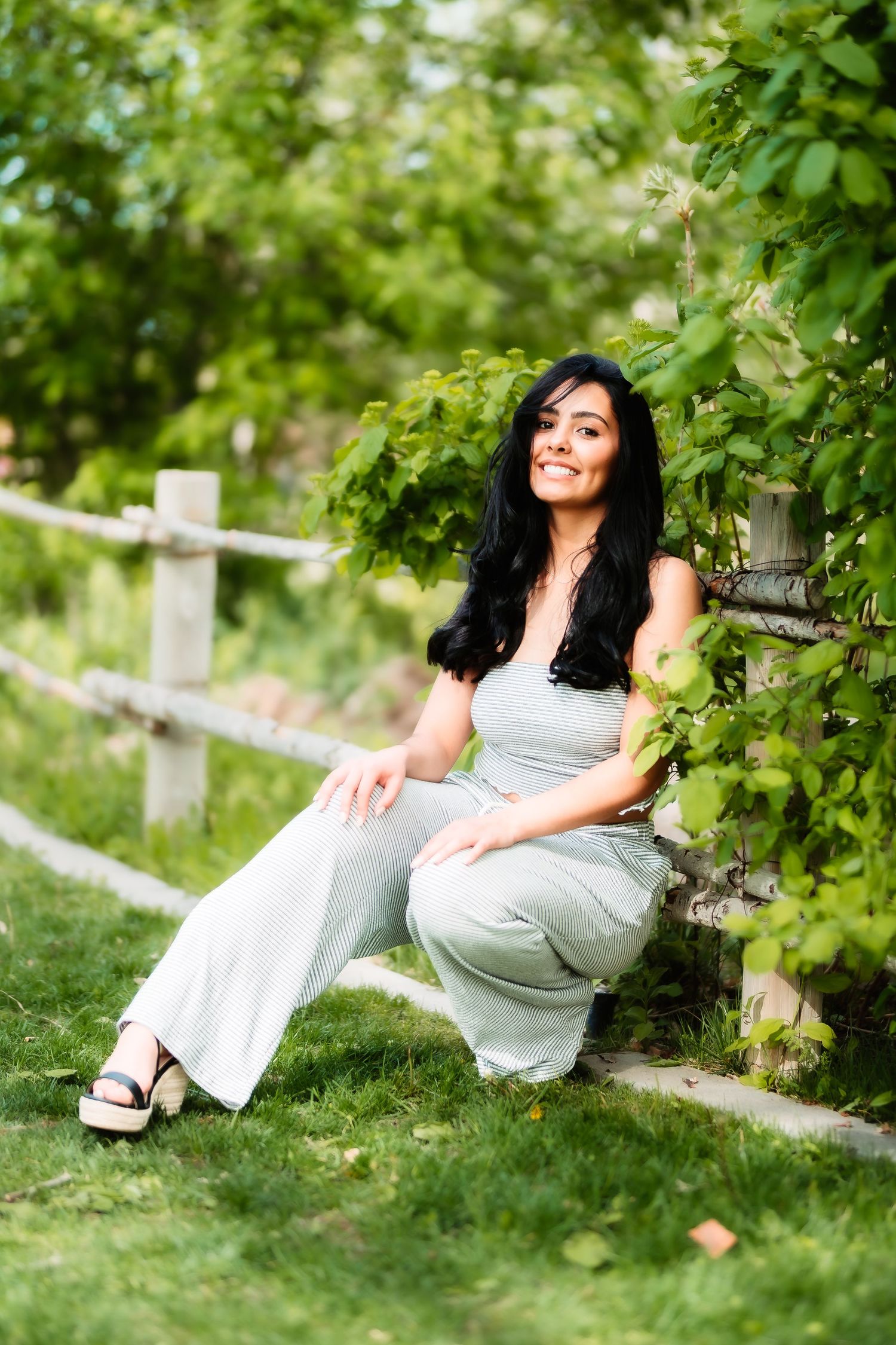 Person in light-colored outfit posing by wooden fence surrounded by green foliage.