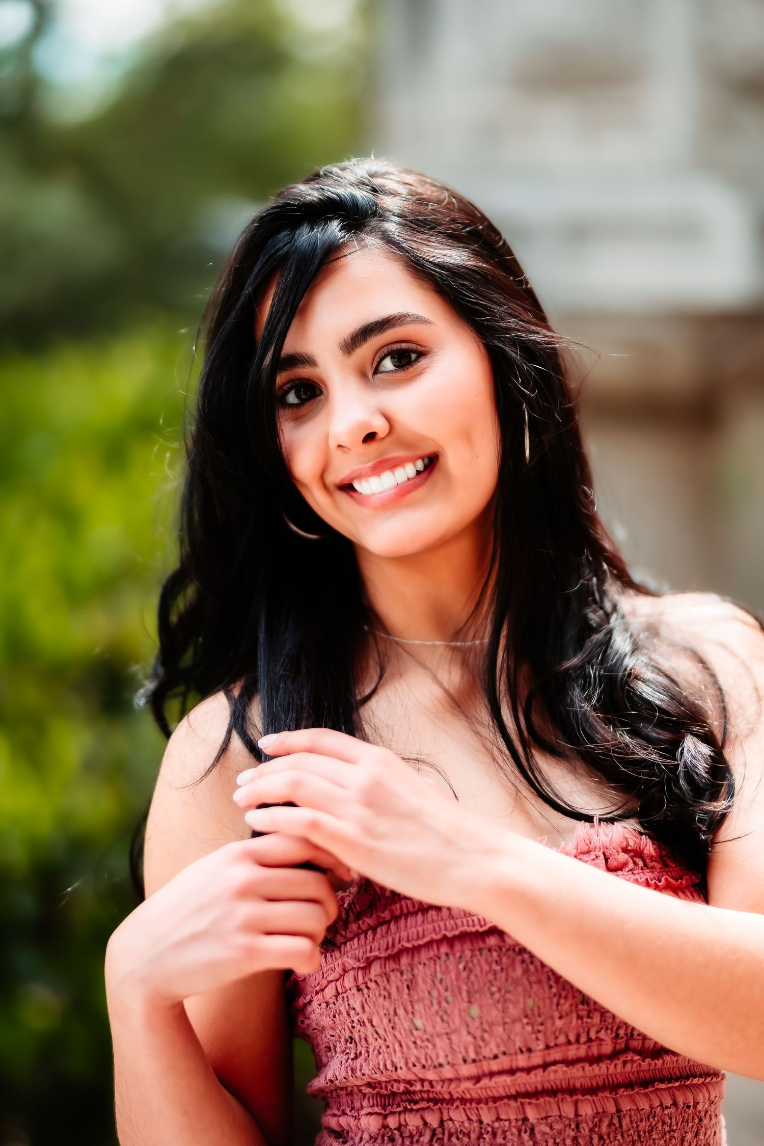 A dark-haired woman in a red smocked dress smiles warmly at the camera in an outdoor setting.