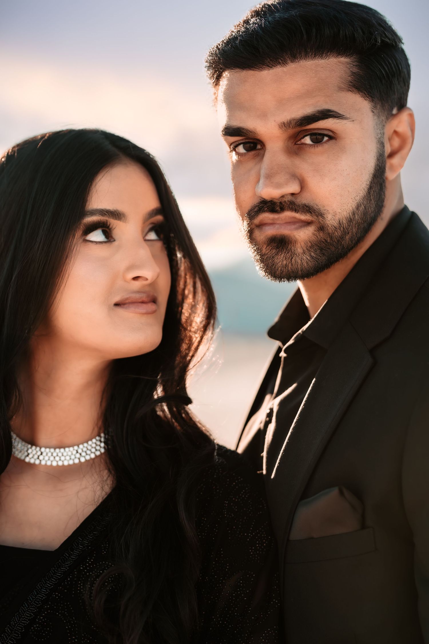 A romantic couple poses in formal wear against a soft background, with her wearing a choker necklace.