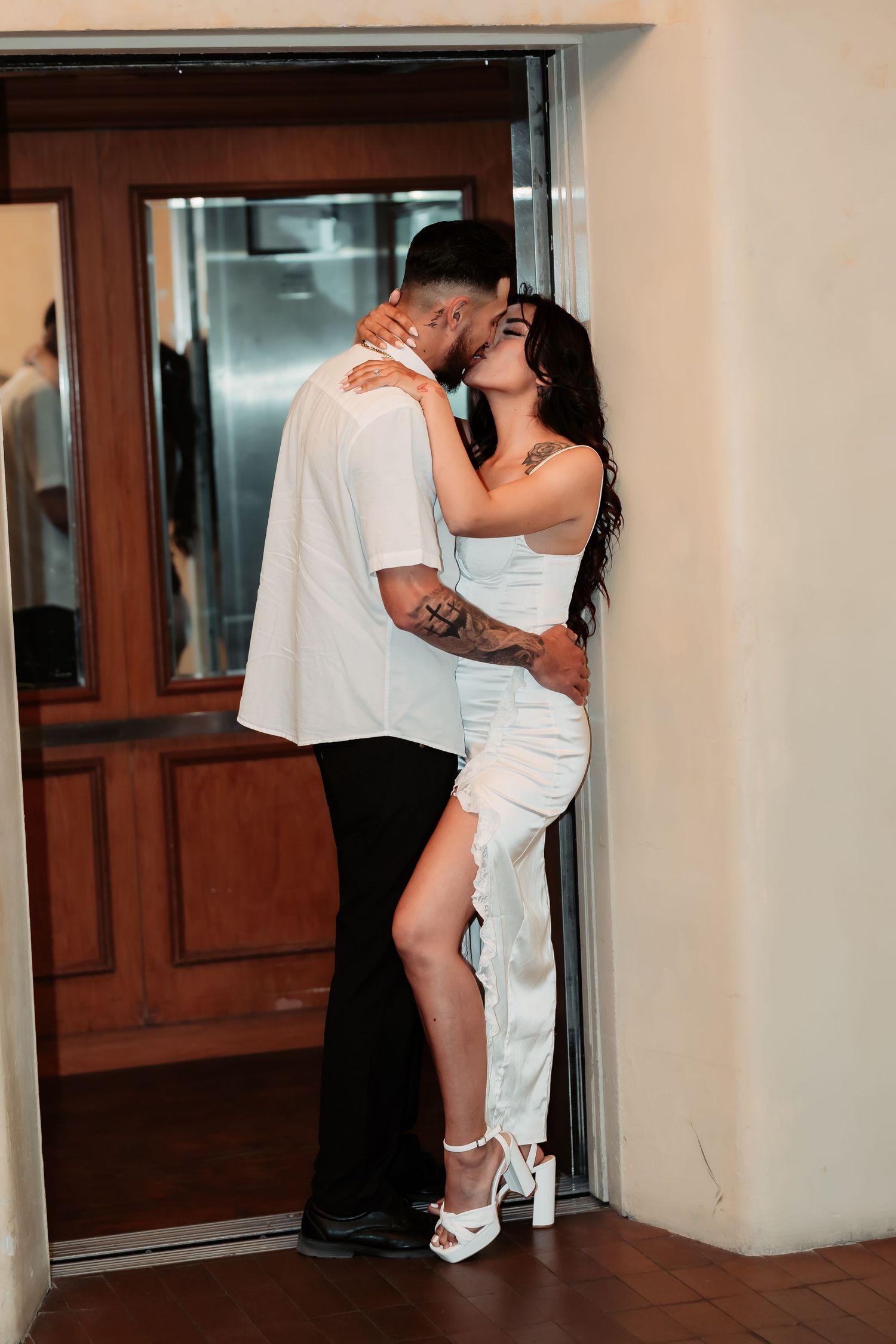 A romantic couple embraces in a doorway, with the woman in white dress lifting her leg while kissing.