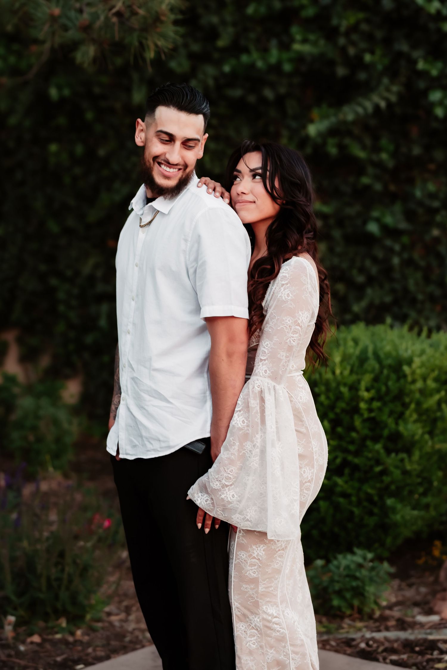 A romantic outdoor portrait shows a stylishly dressed couple in white standing close together among garden foliage.
