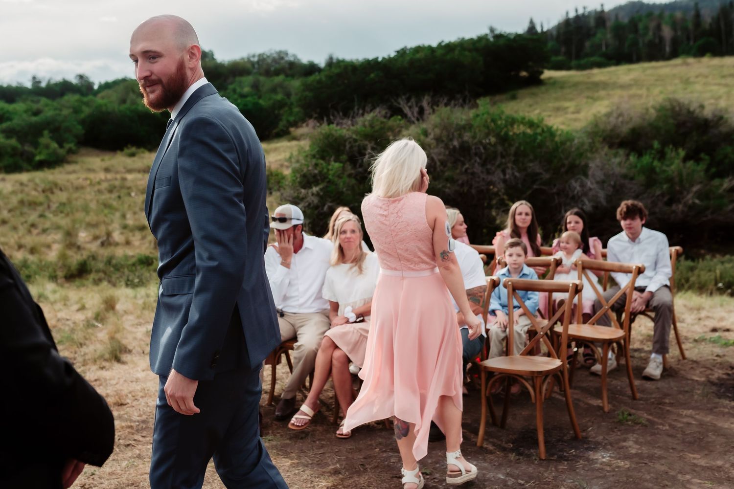 Wedding ceremony setup with wooden chairs and guests in pink dresses against a mountain backdrop.