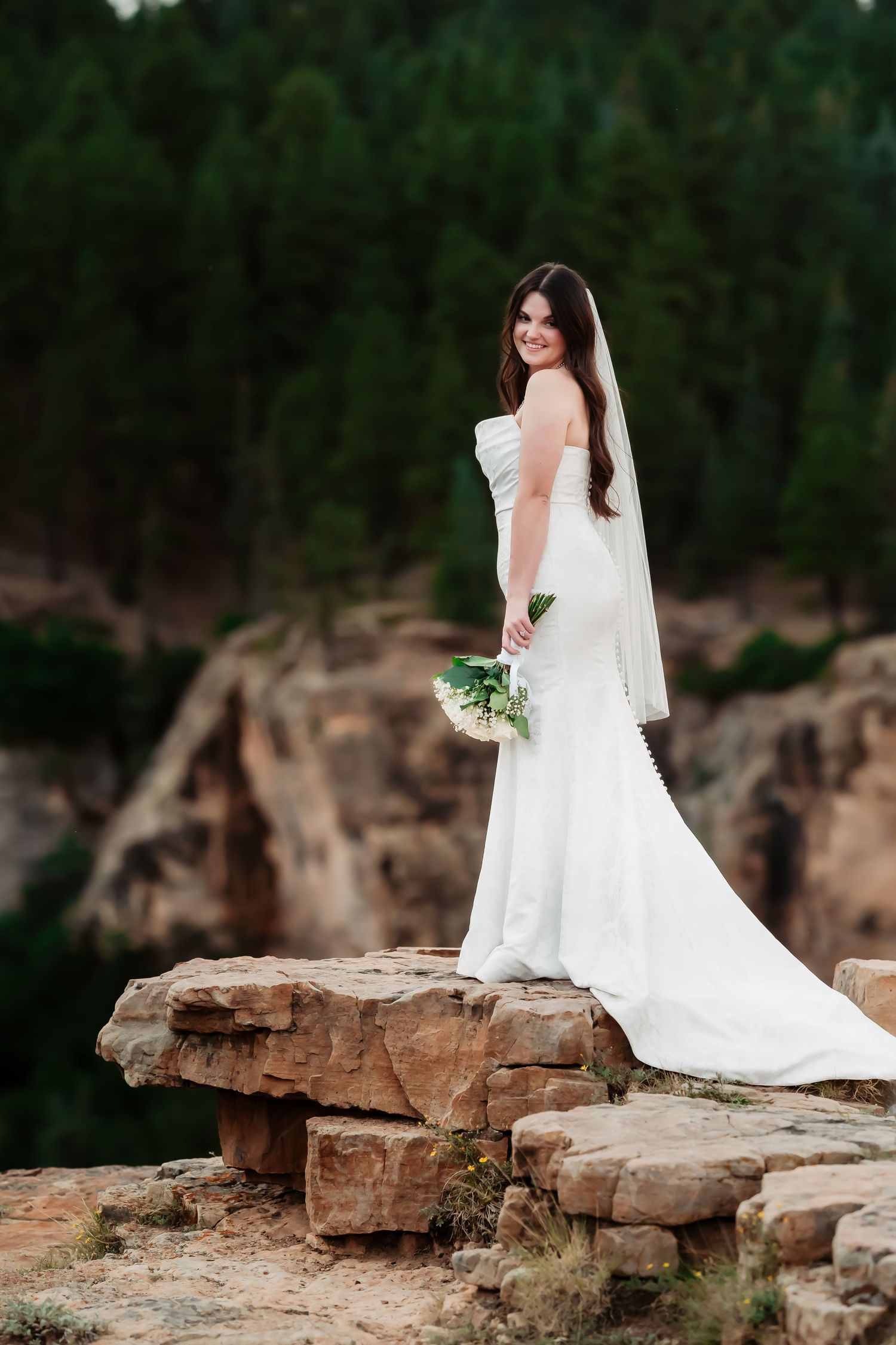 Bride in flowing white dress poses elegantly on rocky mountain ledge.