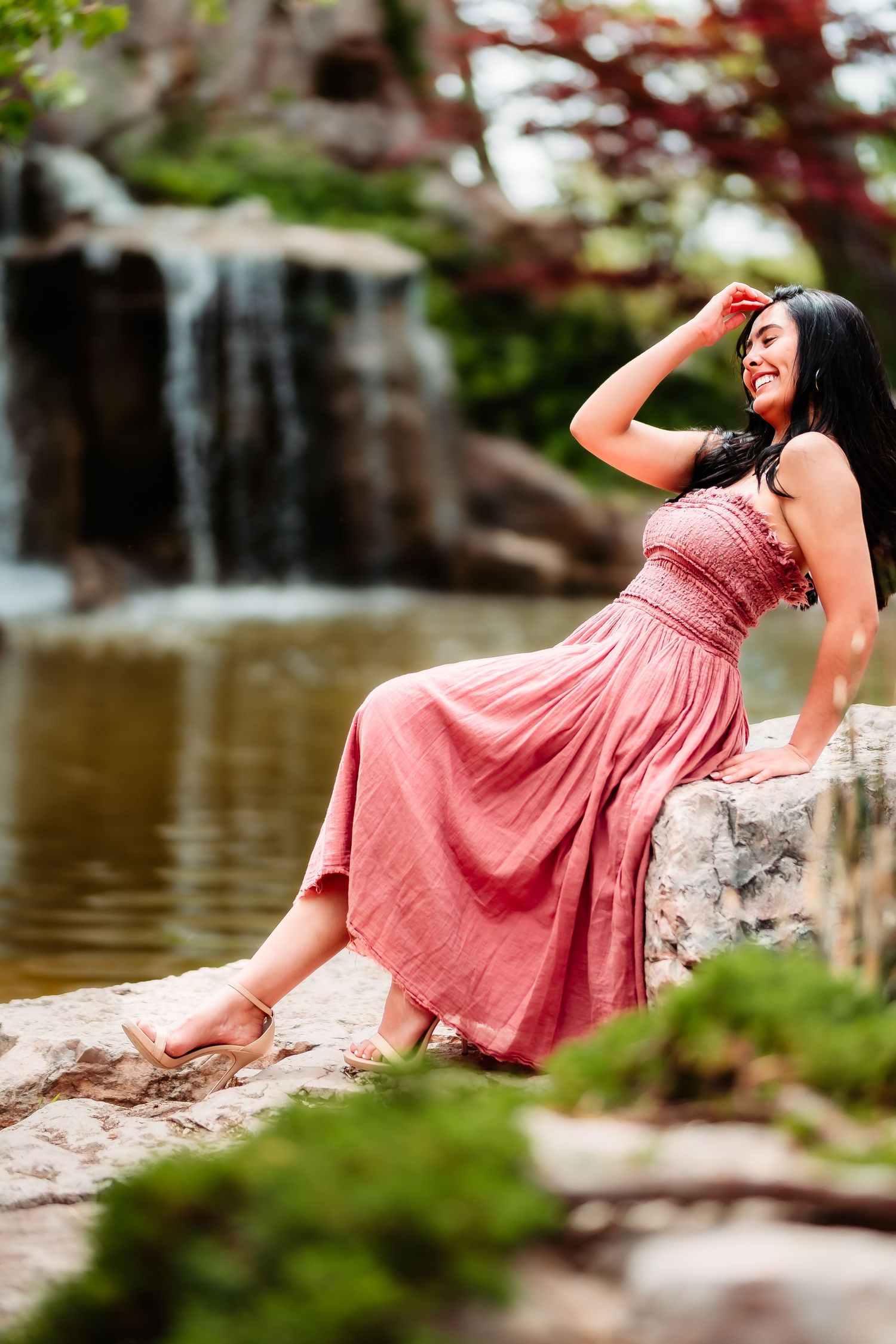 Woman in pink dress posing playfully by waterfall on rock formation.
