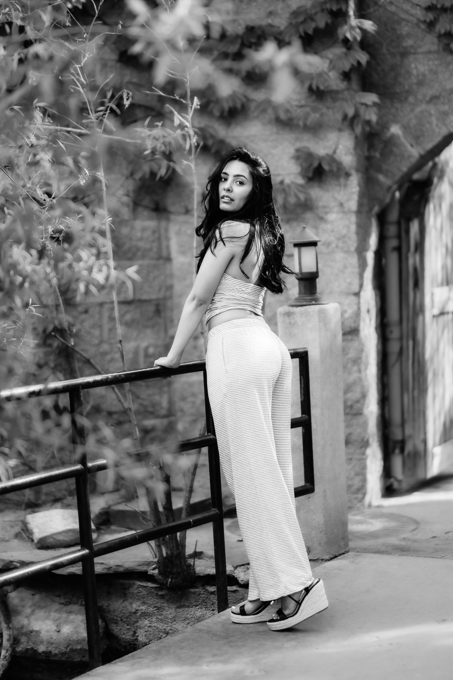 A black and white photo of someone in white attire leaning against a railing with foliage in background.
