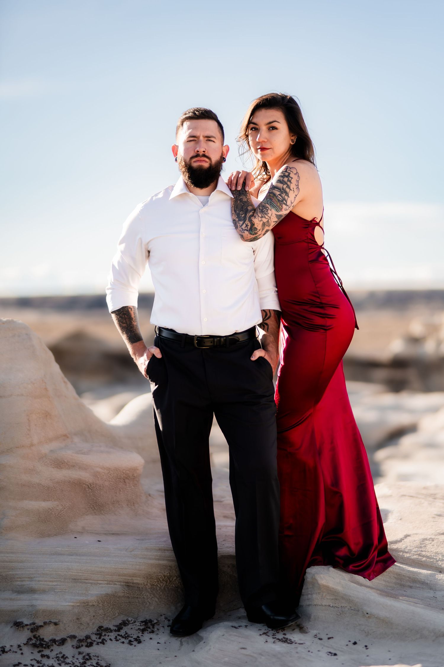 A couple poses together on rocky terrain, with her wearing a flowing red gown and him in formal attire.