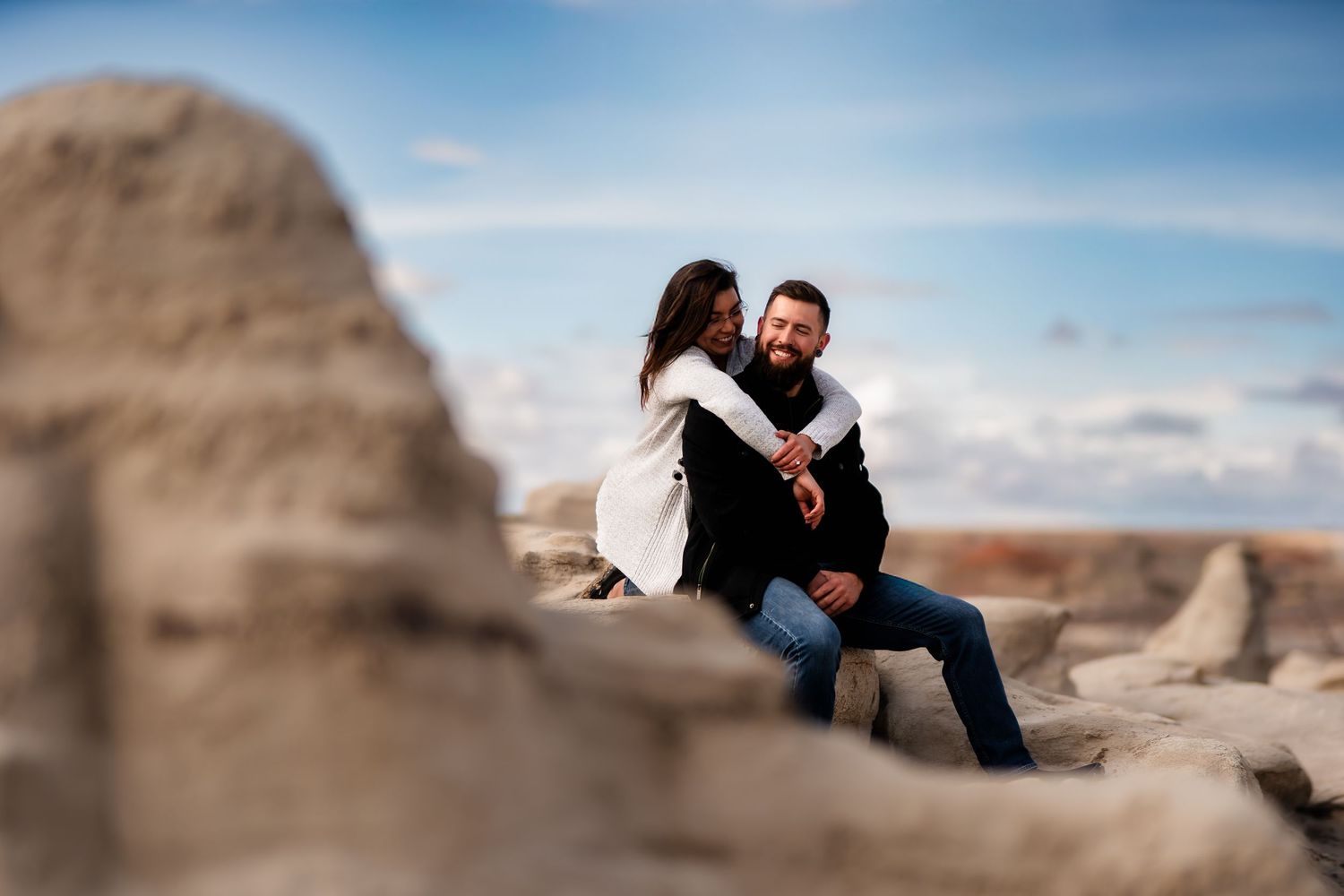 Two people sit close together on a rocky outcrop against a bright blue sky in a desert landscape.