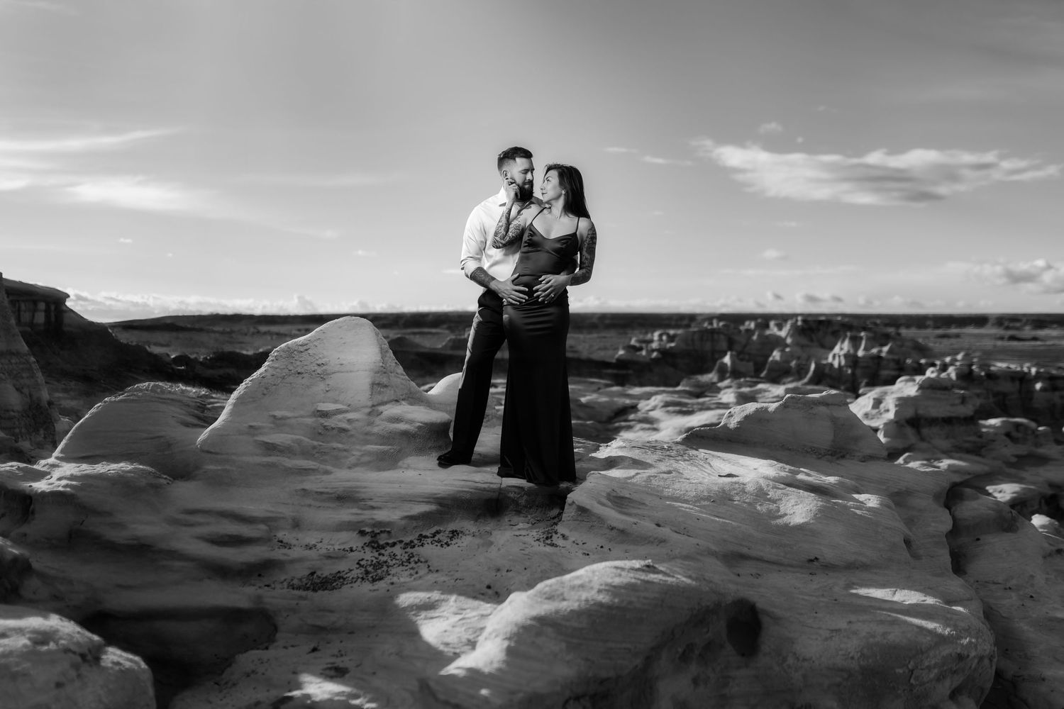 A black and white photo captures a couple standing close together on dramatic desert rock formations.