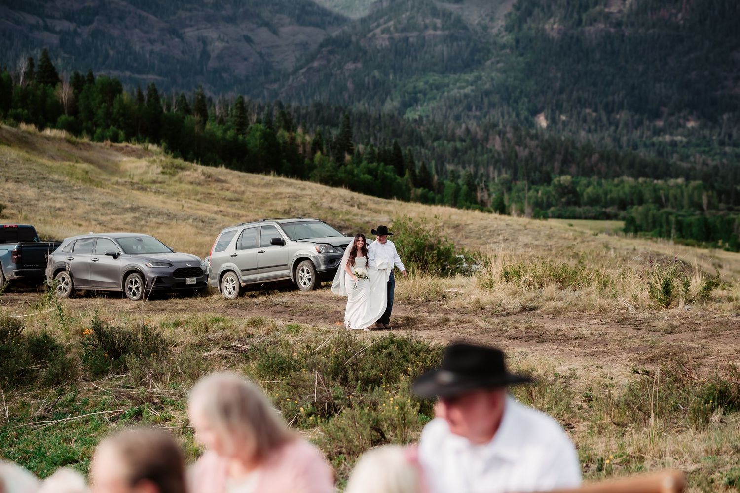 Wide landscape shot of a mountain wedding venue with parked cars in the distance.