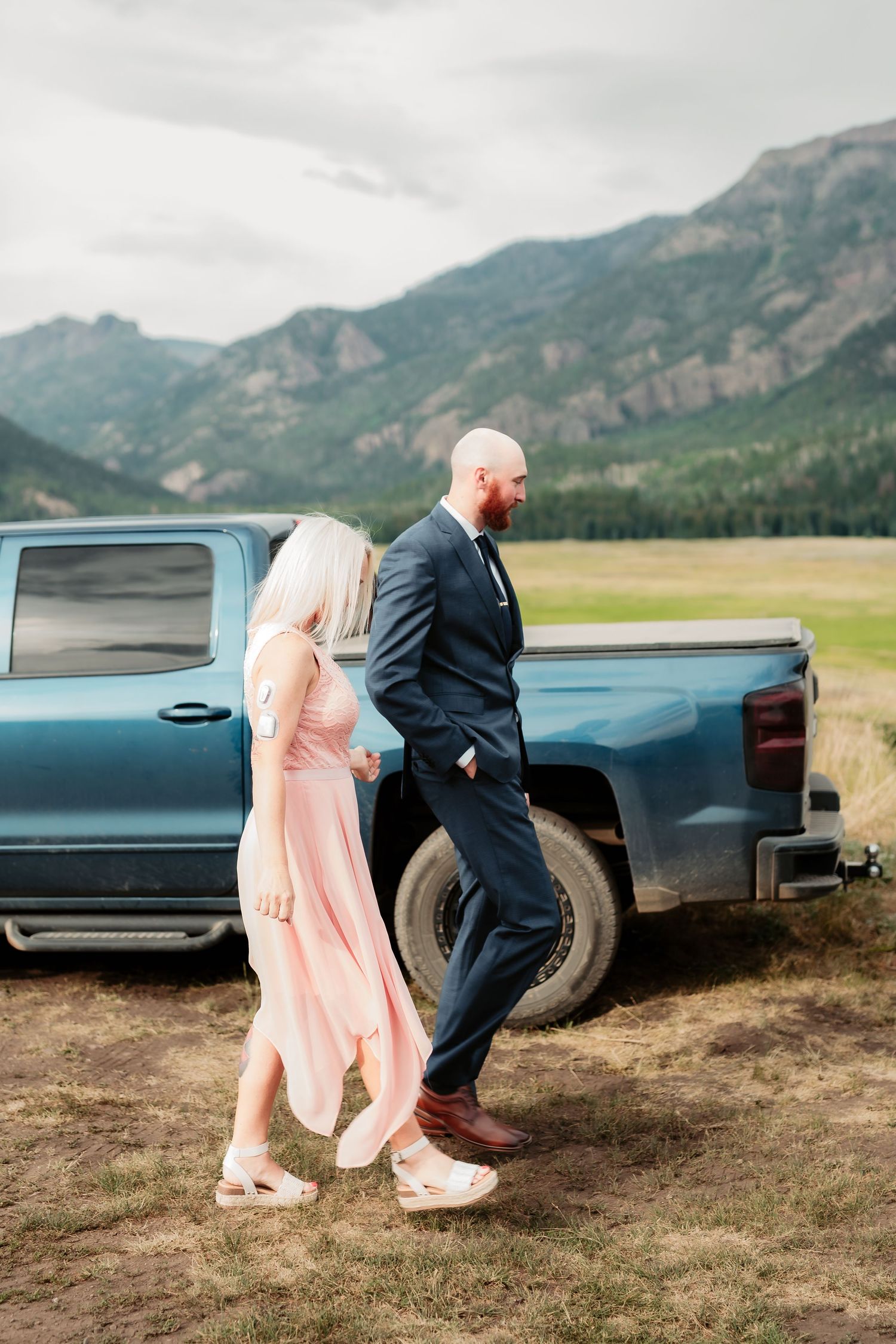A couple walks alongside a pickup truck in matching formal attire against a mountainous landscape.