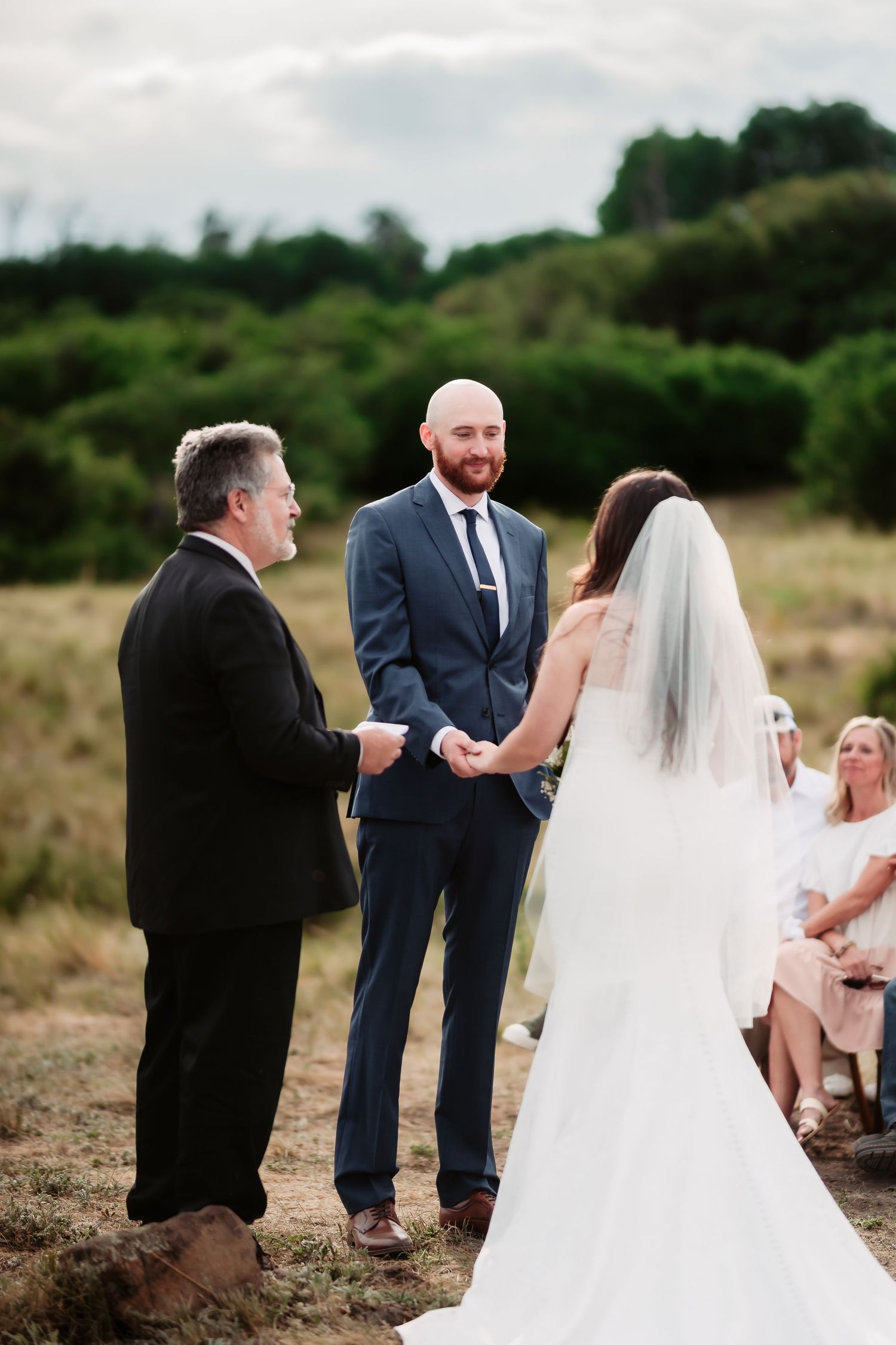 An outdoor wedding ceremony takes place on a hilltop with guests watching as a couple exchanges vows before an officiant.