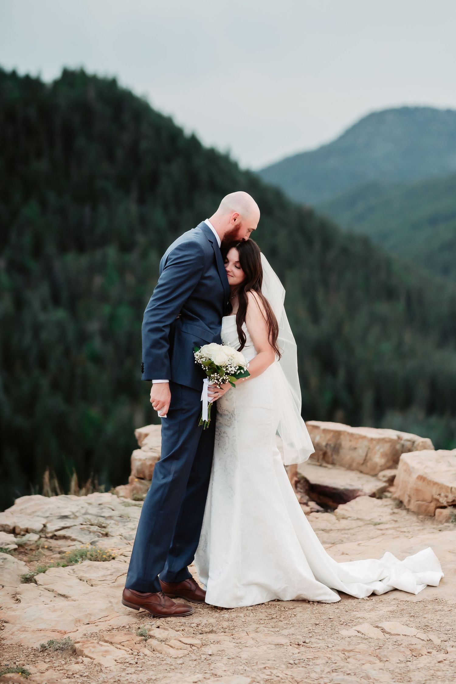 A couple in wedding attire shares a romantic moment on a mountain overlook with evergreen forests in the background.