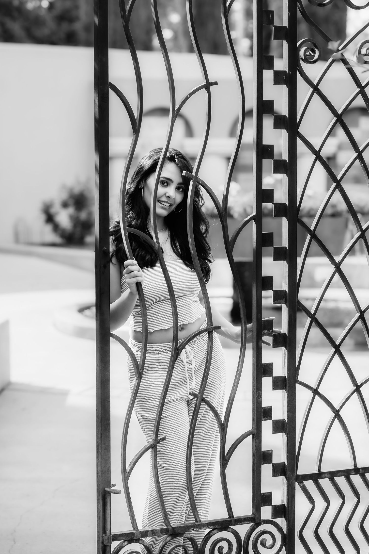 A black and white photo showing a woman framed by ornate metalwork of a garden gate.