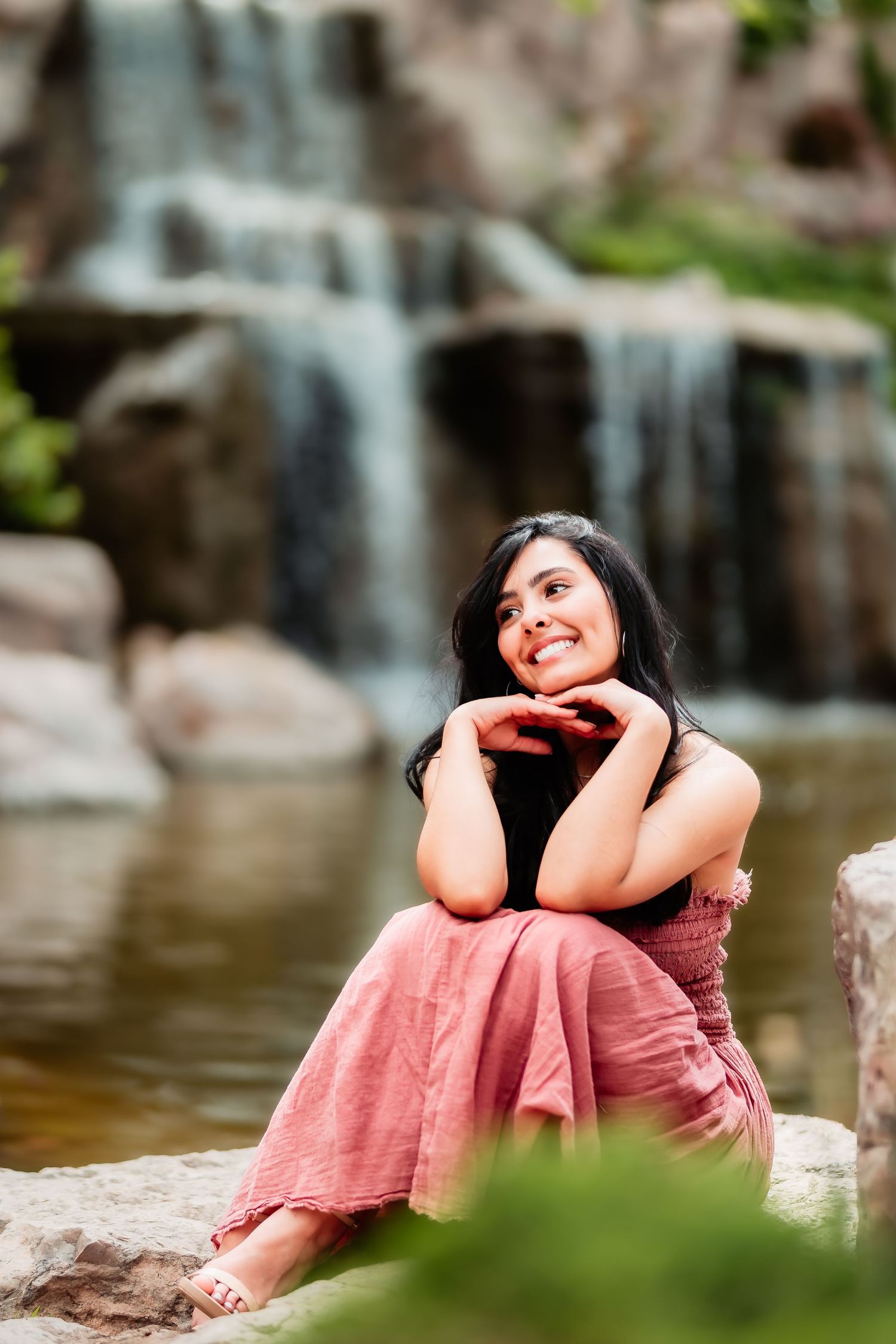 Person in coral dress sits dreamily by waterfall in nature setting with blurred cascade in background.