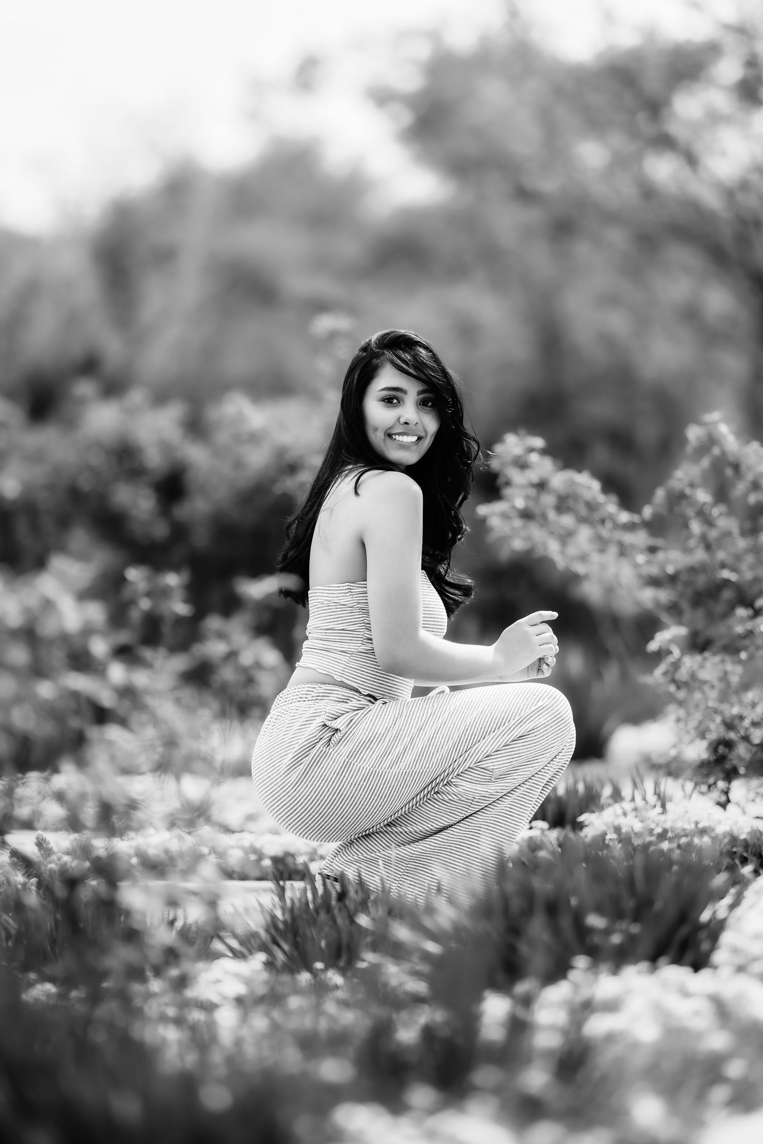 Black and white image of someone crouching among garden flowers in casual attire.
