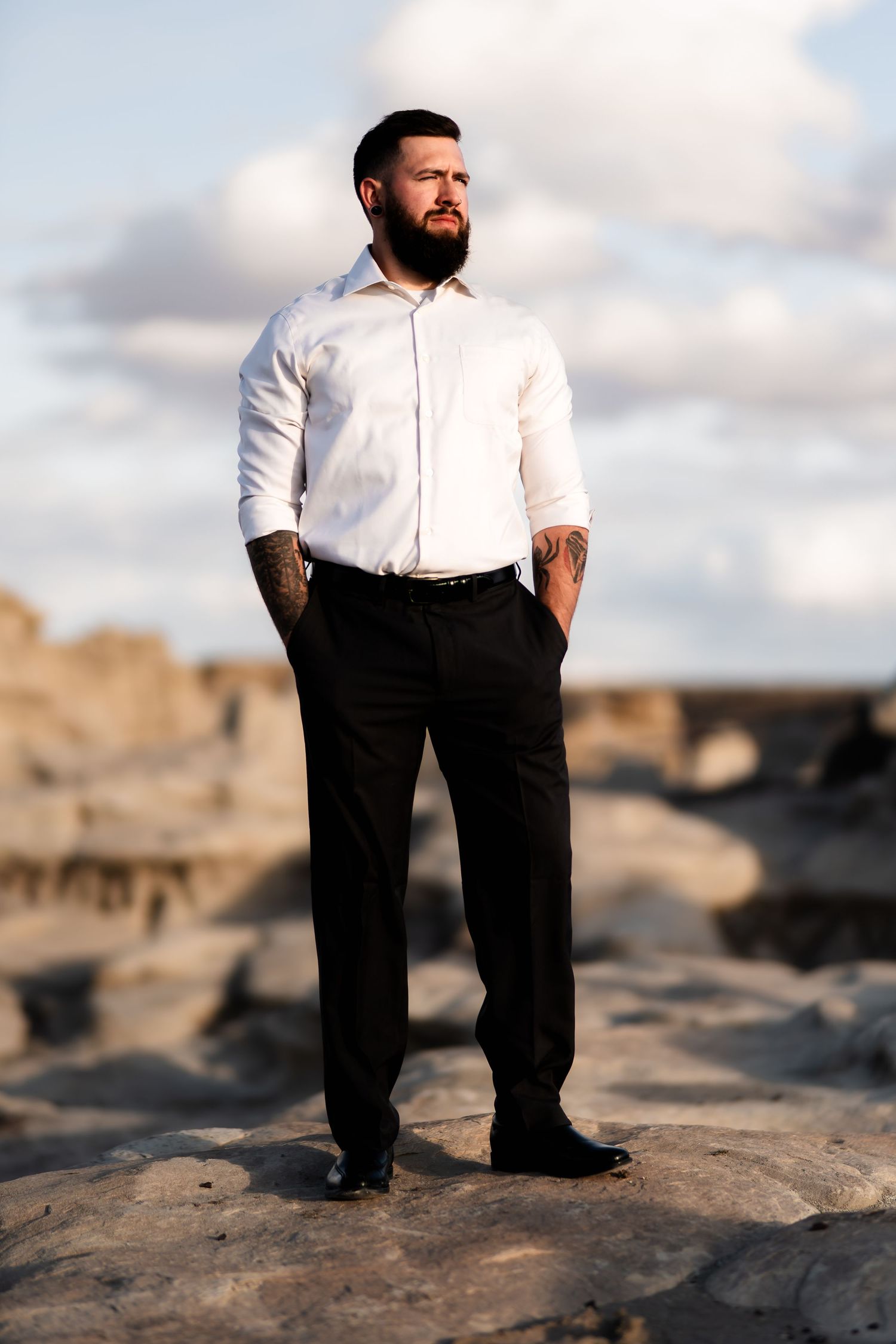 Person in formal attire stands confidently on rocky terrain against cloudy sky.