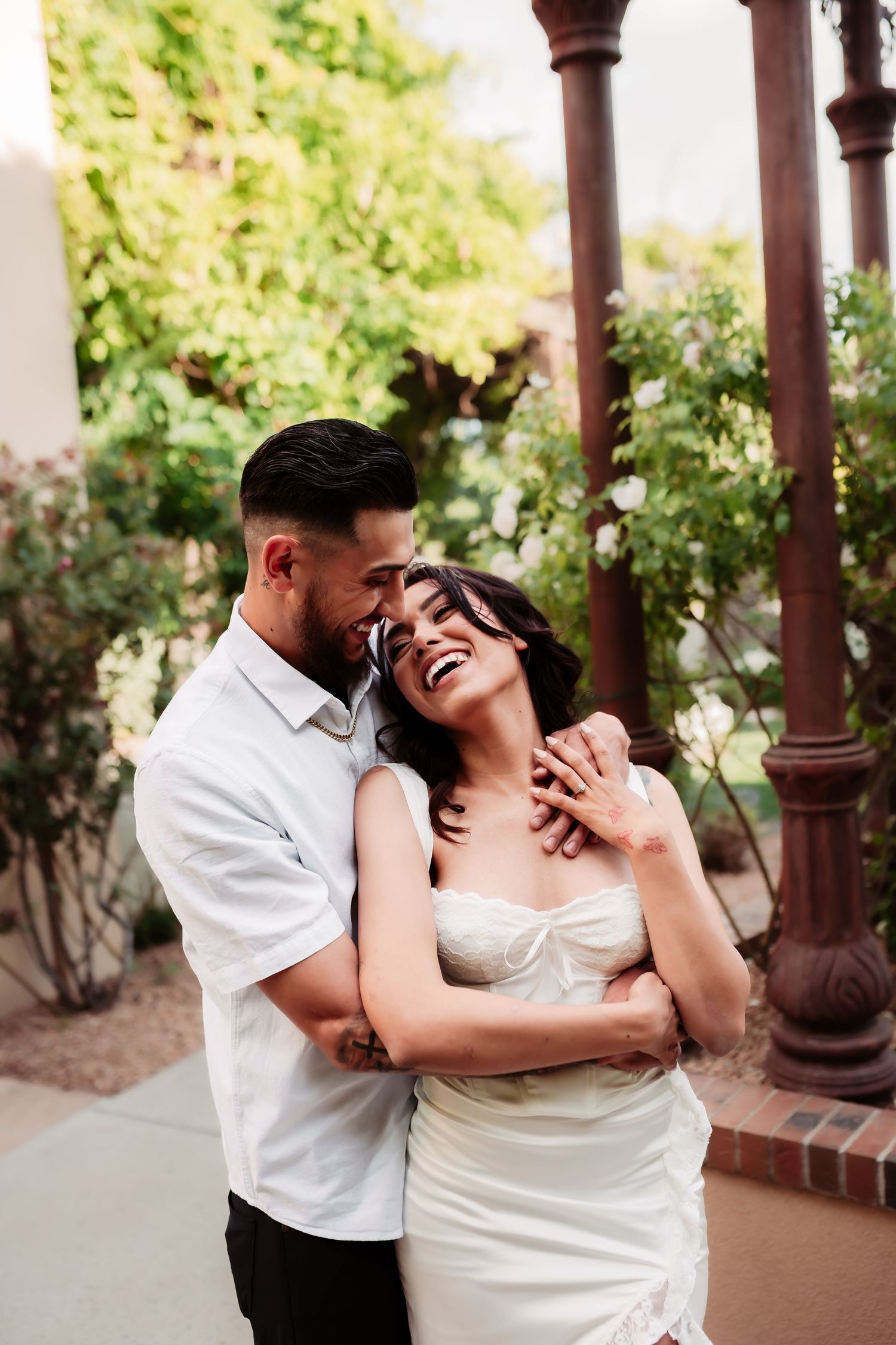 A romantic couple embraces and laughs together under wooden pillars in an outdoor garden setting.