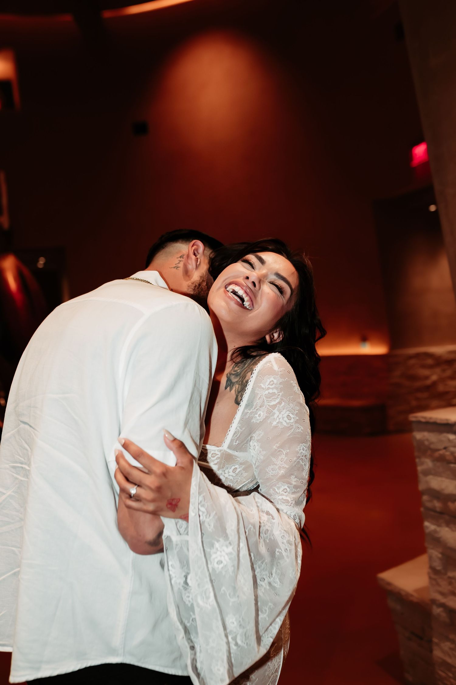 Joyful couple embracing and laughing in dimly lit room with warm red lighting.