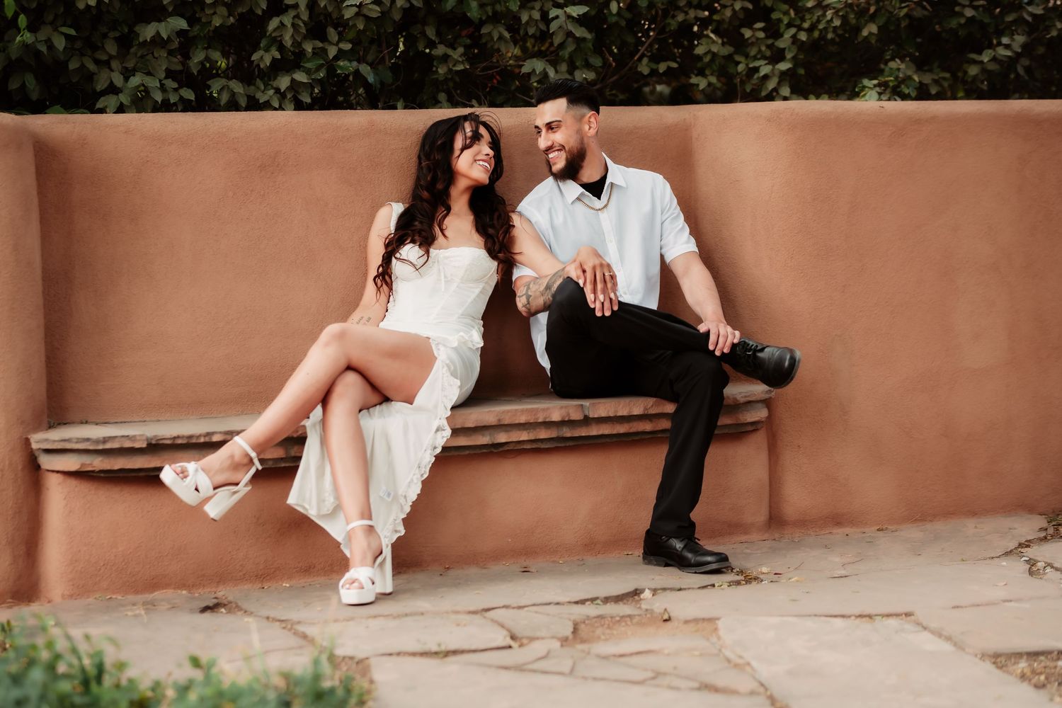 Romantic couple in white sitting on adobe wall during outdoor photoshoot.