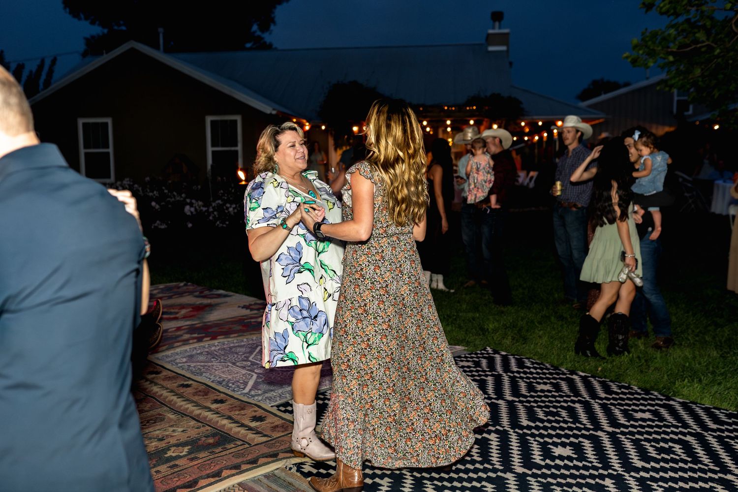 Evening outdoor reception with guests dancing on patterned rug under string lights.