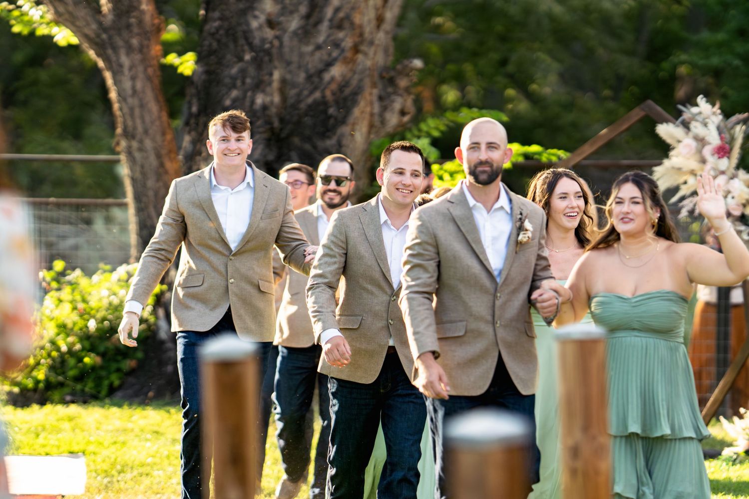 Wedding party members walk together wearing coordinated tan suits and sage green dresses.