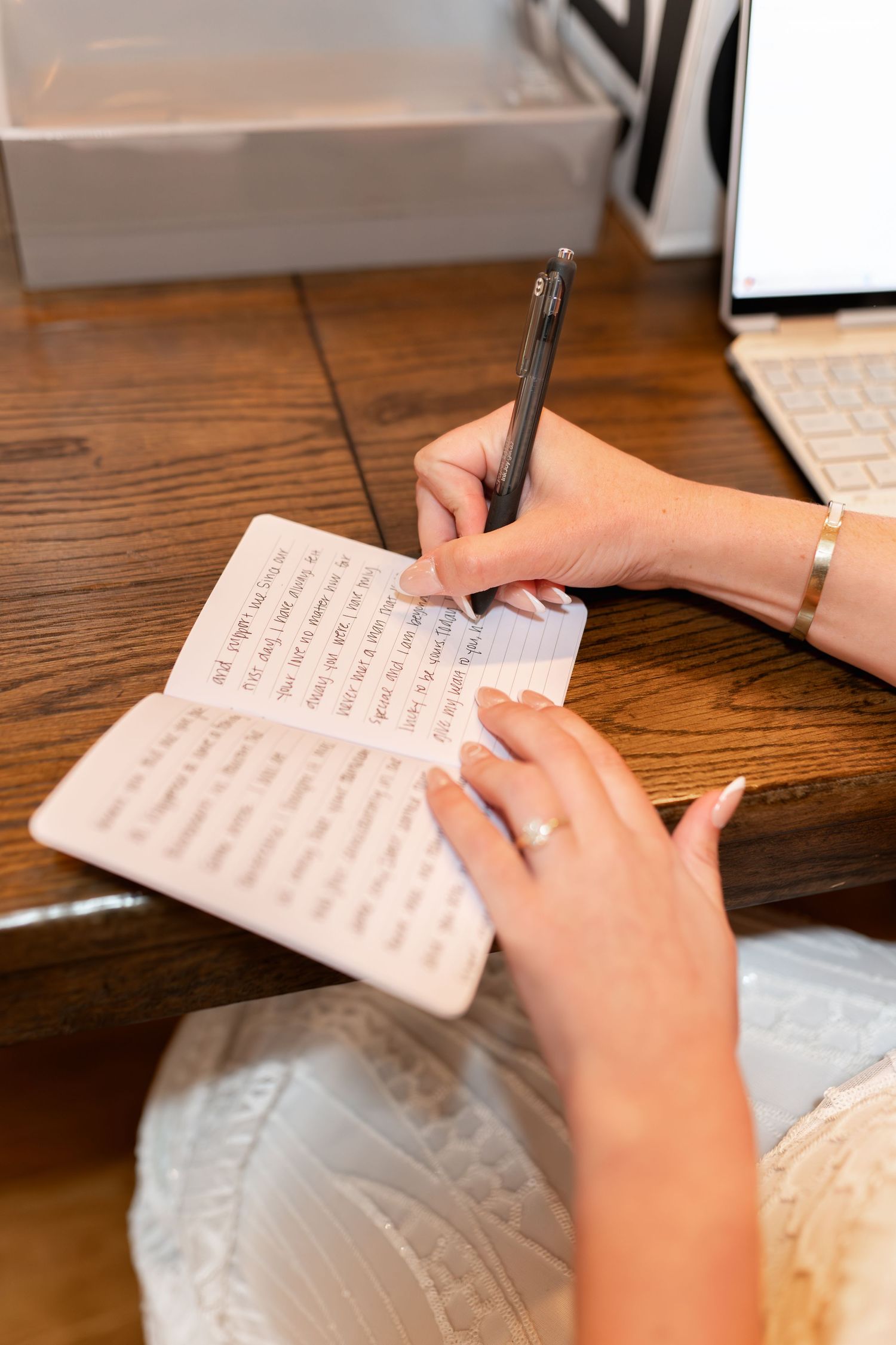A person's hands writing in a notebook with a pen on a wooden desk next to a computer keyboard.