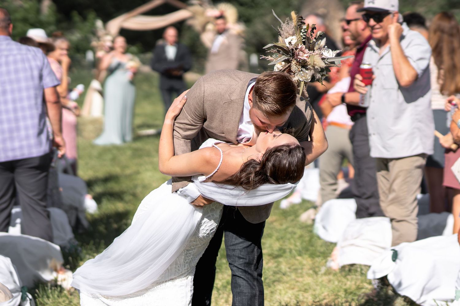 A romantic outdoor wedding moment as a couple shares a dip kiss on their special day with guests looking on.