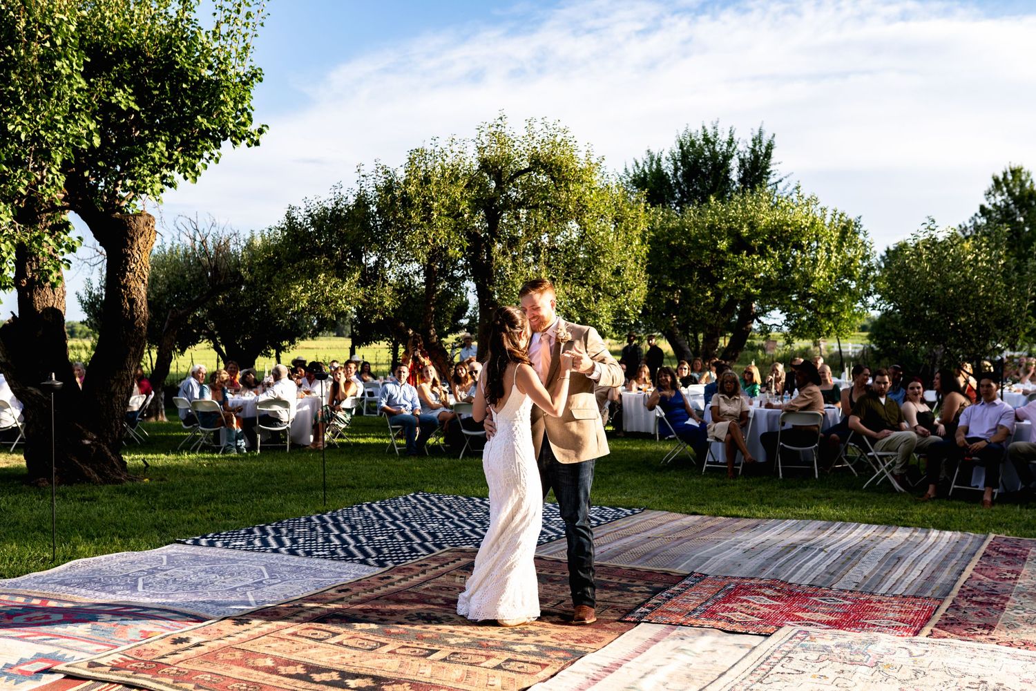 Newlyweds share their first dance on a patterned outdoor dance floor surrounded by trees and guests.
