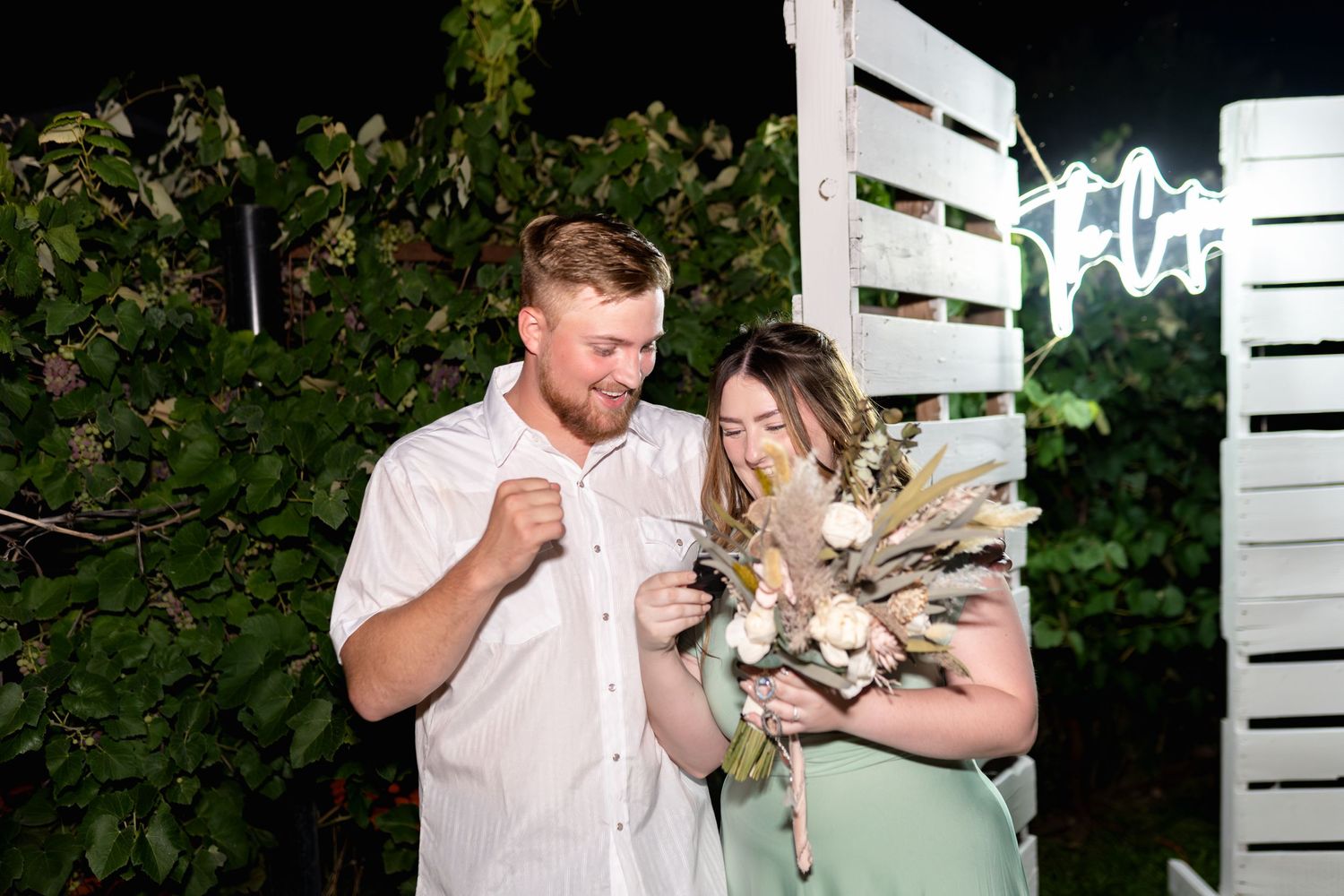 Couple shares playful moment at night against white wooden backdrop with ambient lighting.